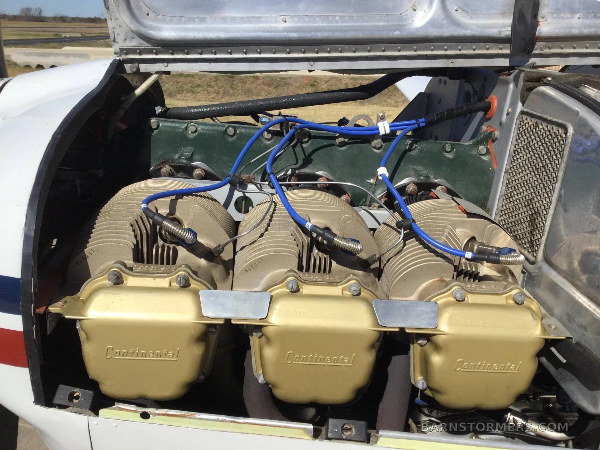 A close-up view of a Continental aircraft engine with three gold valve covers and blue ignition wires, with the cowling open.