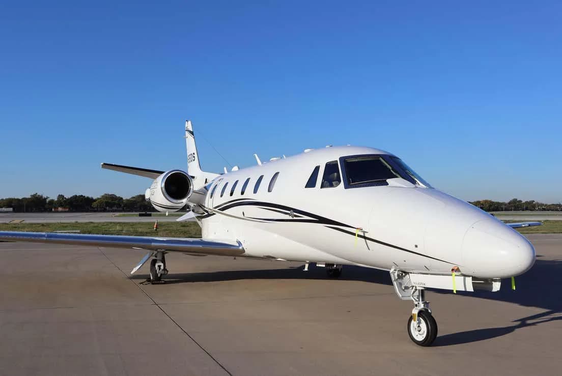 A white business jet with black and gold accent stripes is parked on an airport tarmac under a clear blue sky, viewed from the front-right.