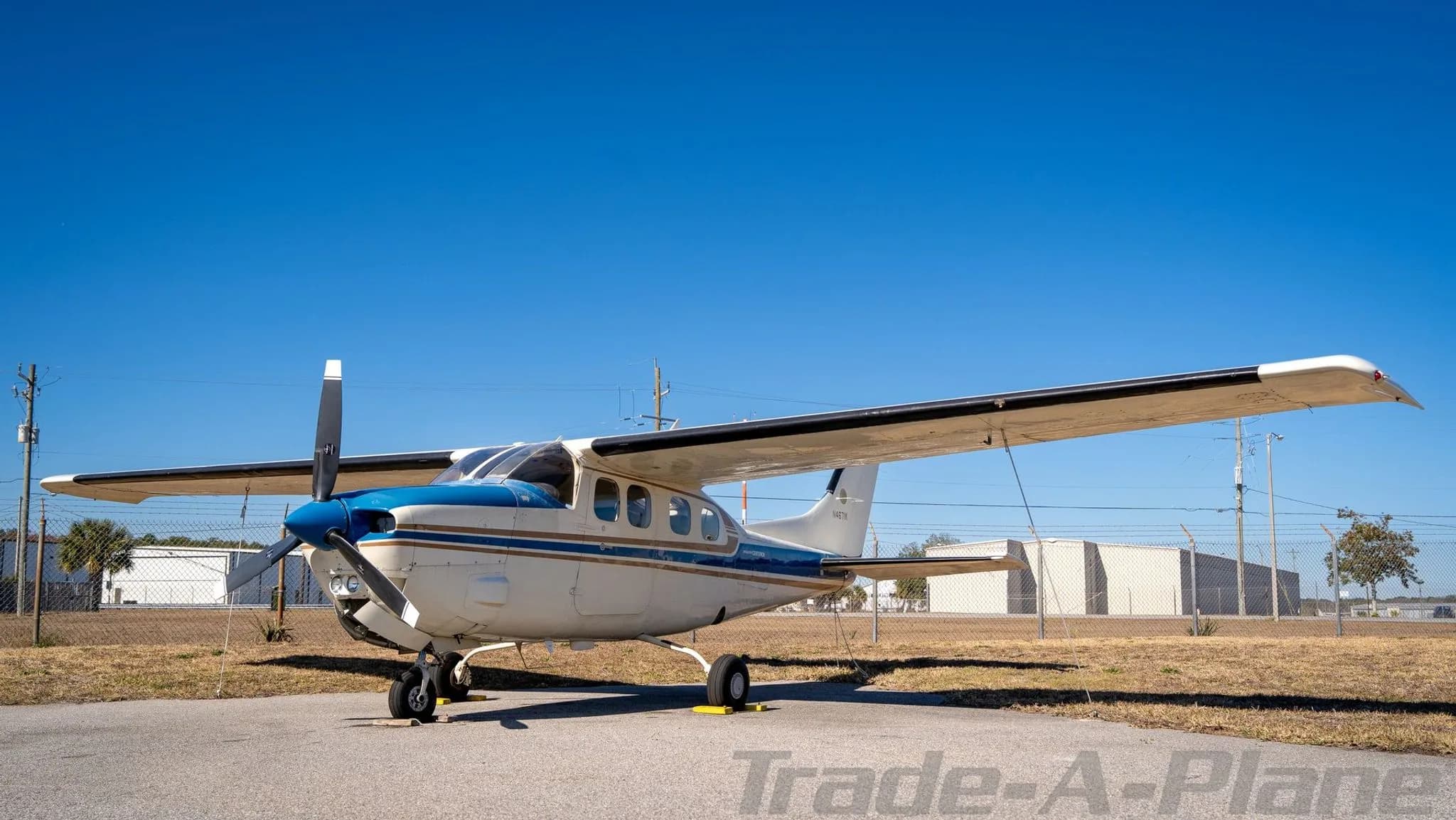 A white and blue Cessna Centurion N4671K aircraft parked on an asphalt ramp under a clear blue sky.