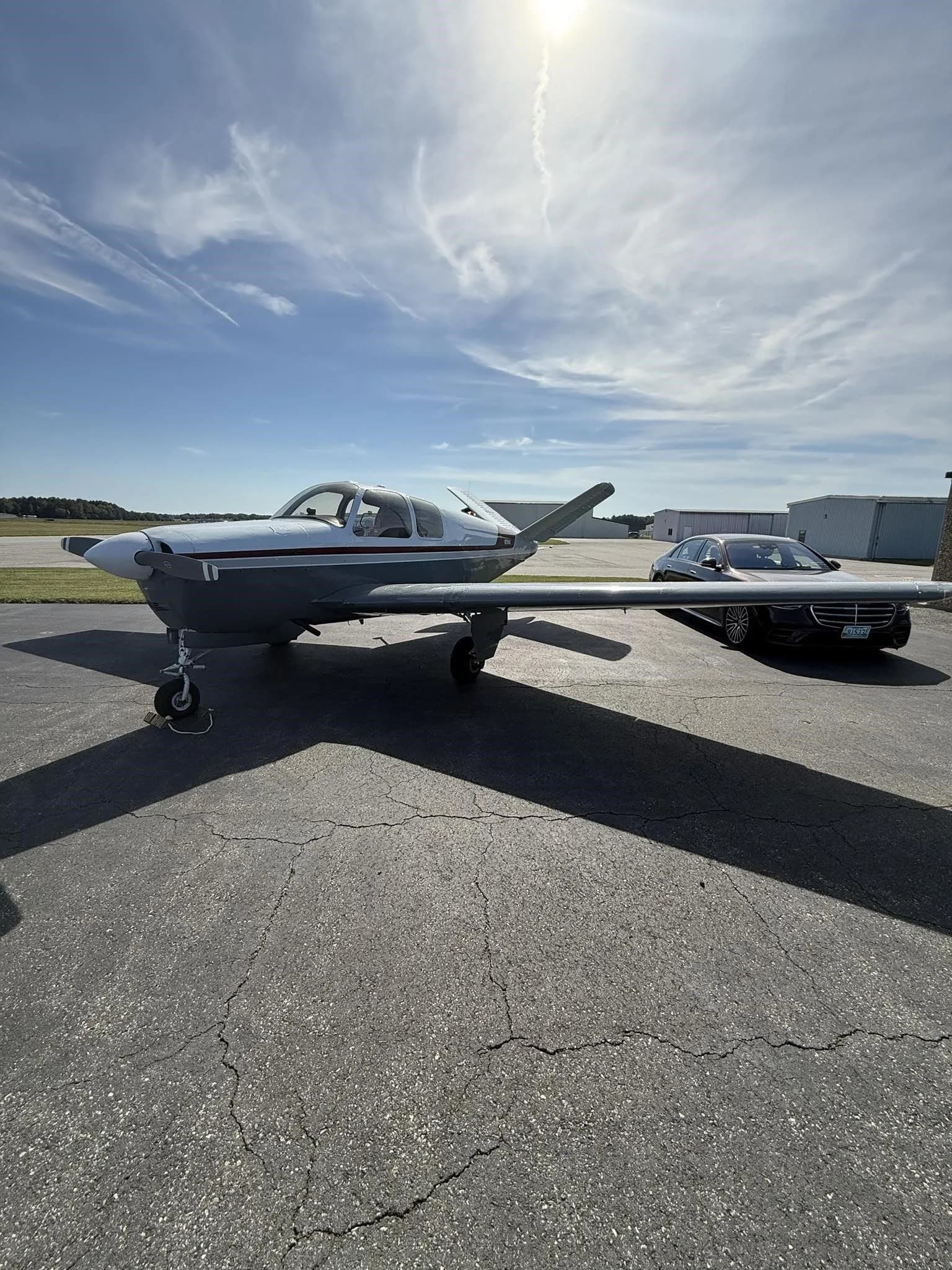 The N2194D Beechcraft Bonanza V-tail aircraft, with a white and grey paint scheme and red stripe, parked on an airfield tarmac with a car in the background.