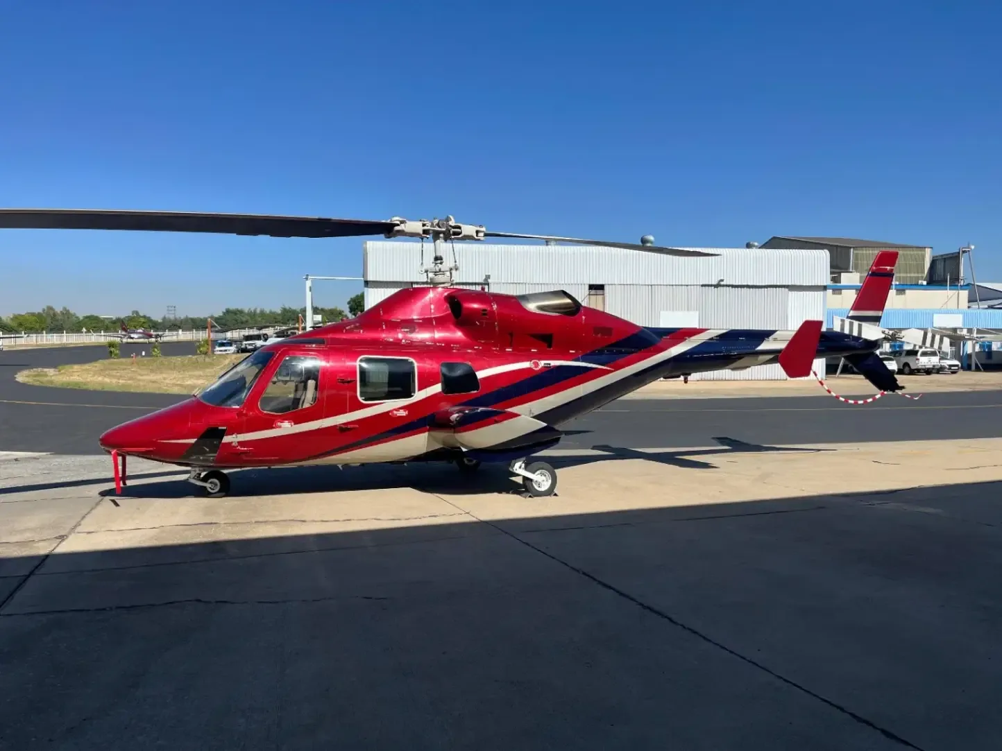 A red and white helicopter sits on the tarmac in front of a hangar.