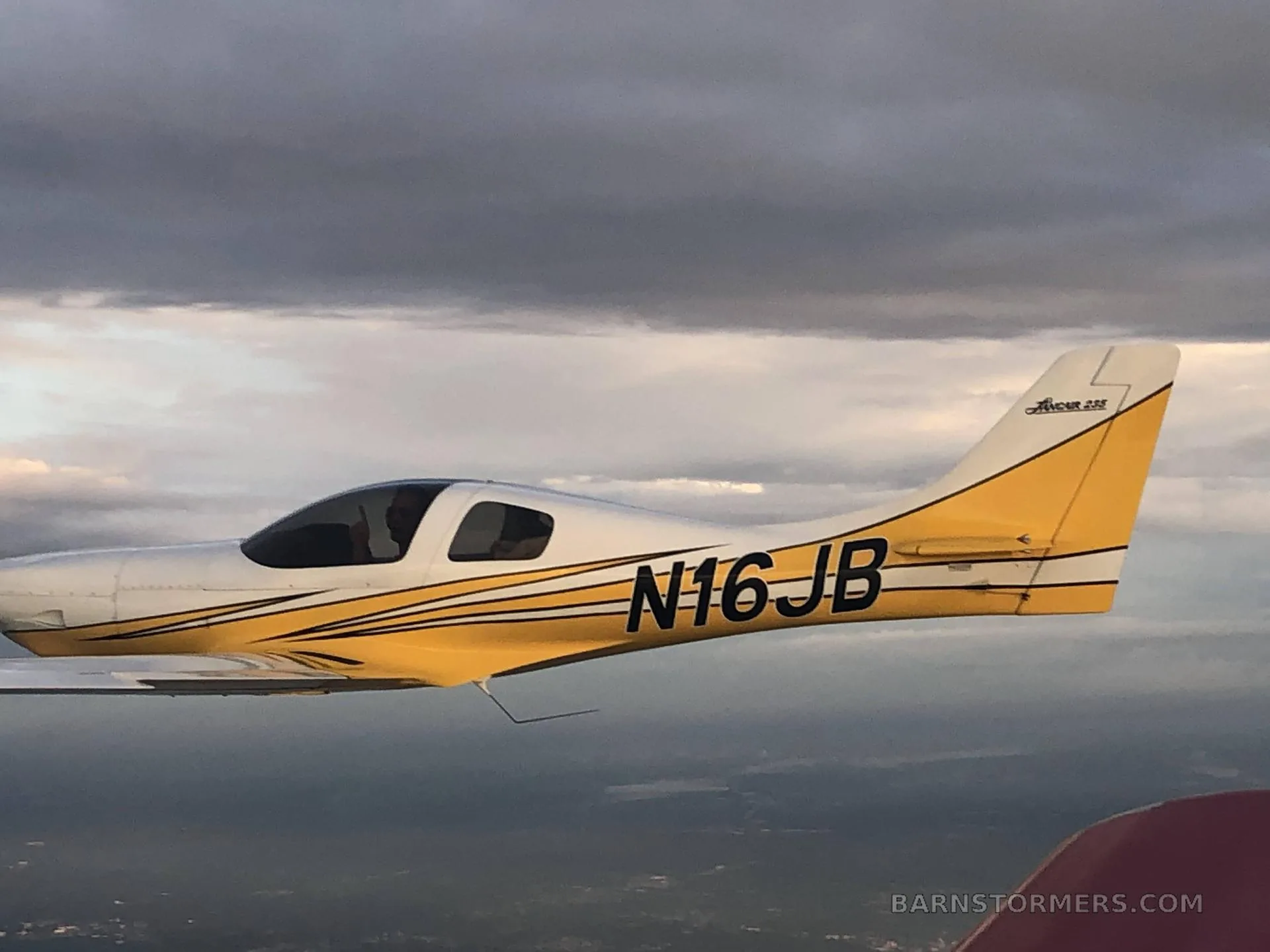A white and yellow single-engine aircraft with tail number N16JB, flying against a cloudy sky.