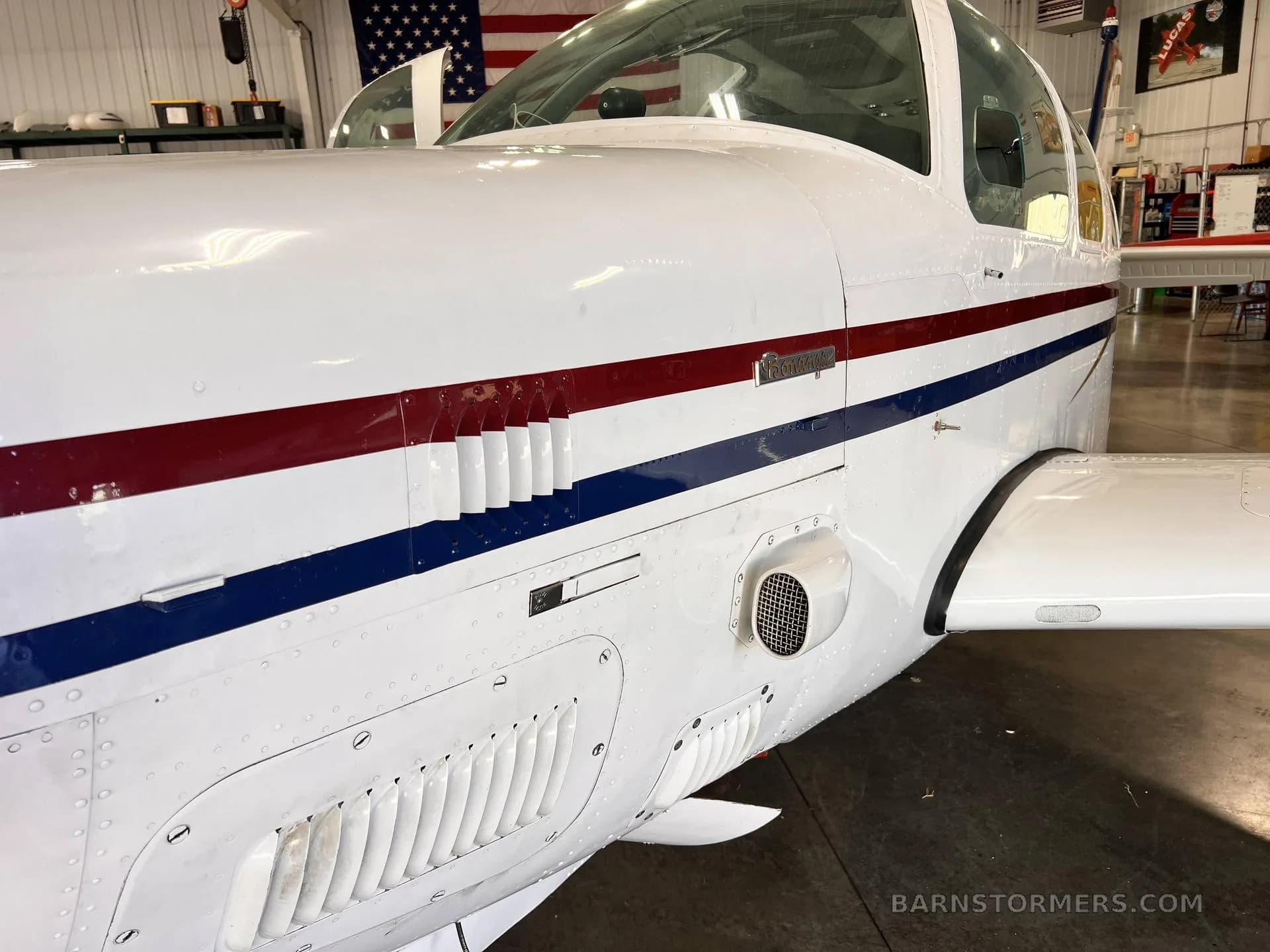 A close-up side view of the white fuselage of a single-engine aircraft with red and blue stripes, showing engine vents.