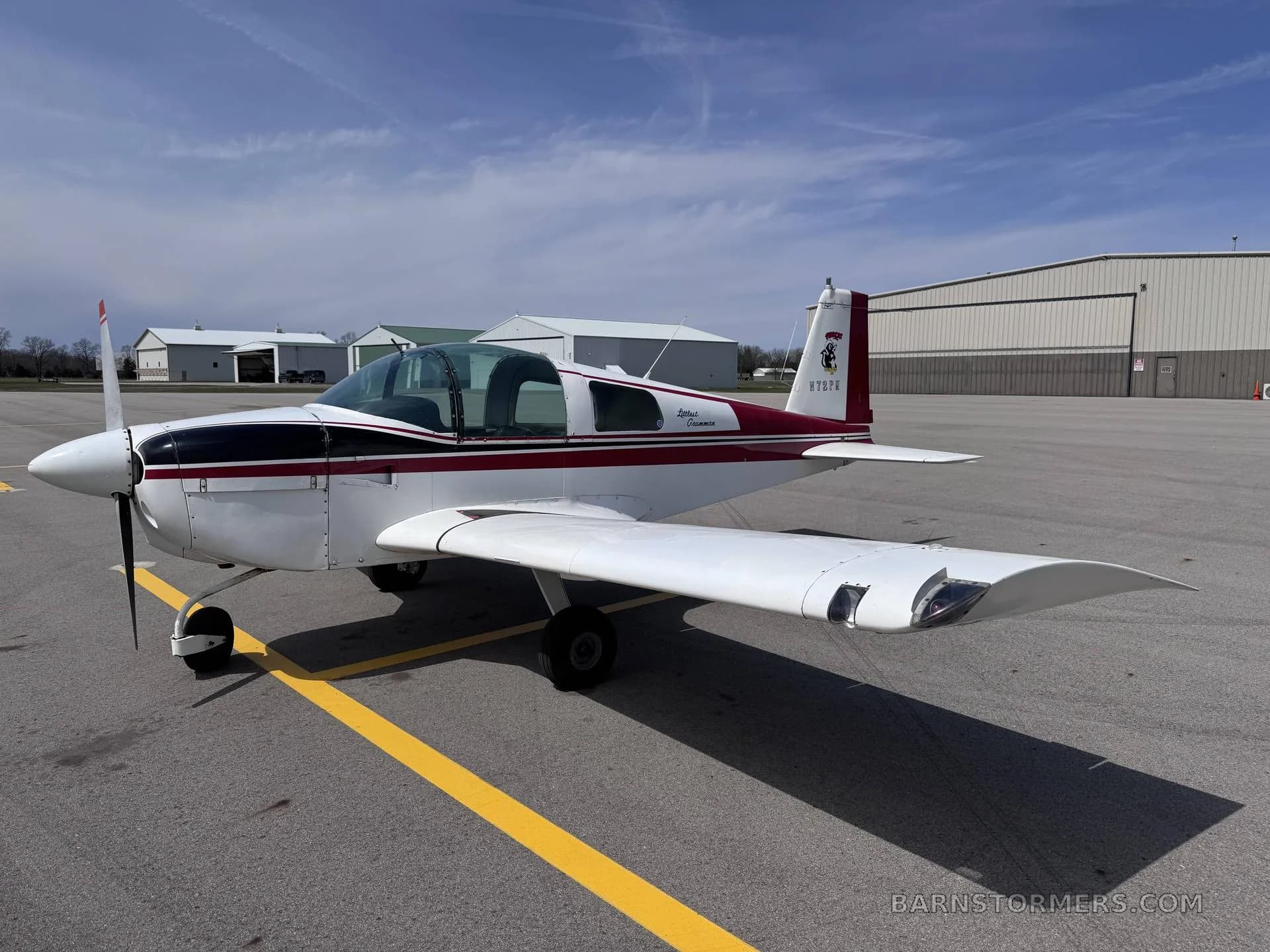 A white and maroon Grumman American AA-5 Traveler aircraft is parked on an asphalt tarmac with hangars in the background.