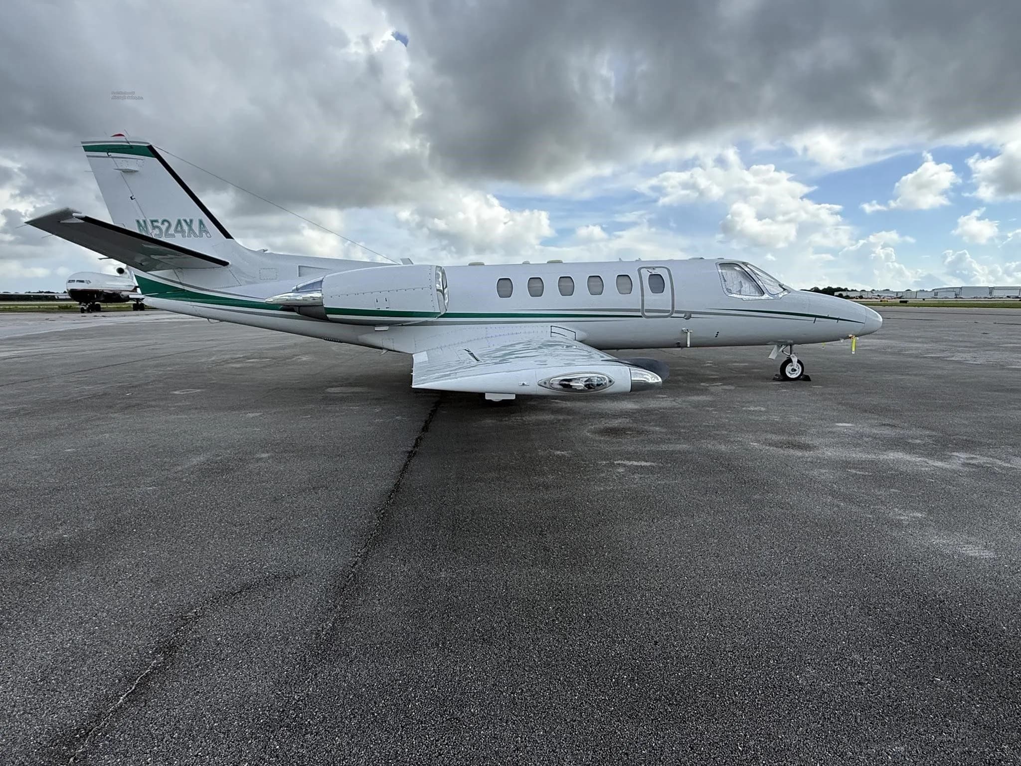 A side view of the white business jet N524XA with green stripes, parked on an airport tarmac under a cloudy sky.