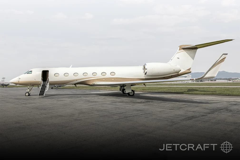 A white and gold Gulfstream G650 business jet with its door open and stairs extended, parked on an asphalt tarmac.