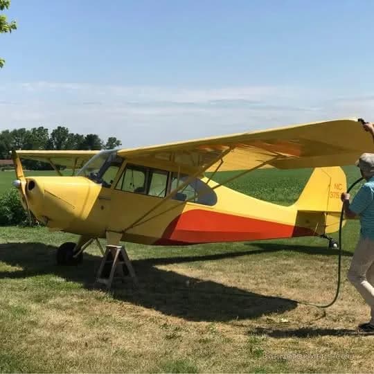 A yellow and orange single-engine high-wing aircraft is parked on grass, with a person hosing it down.