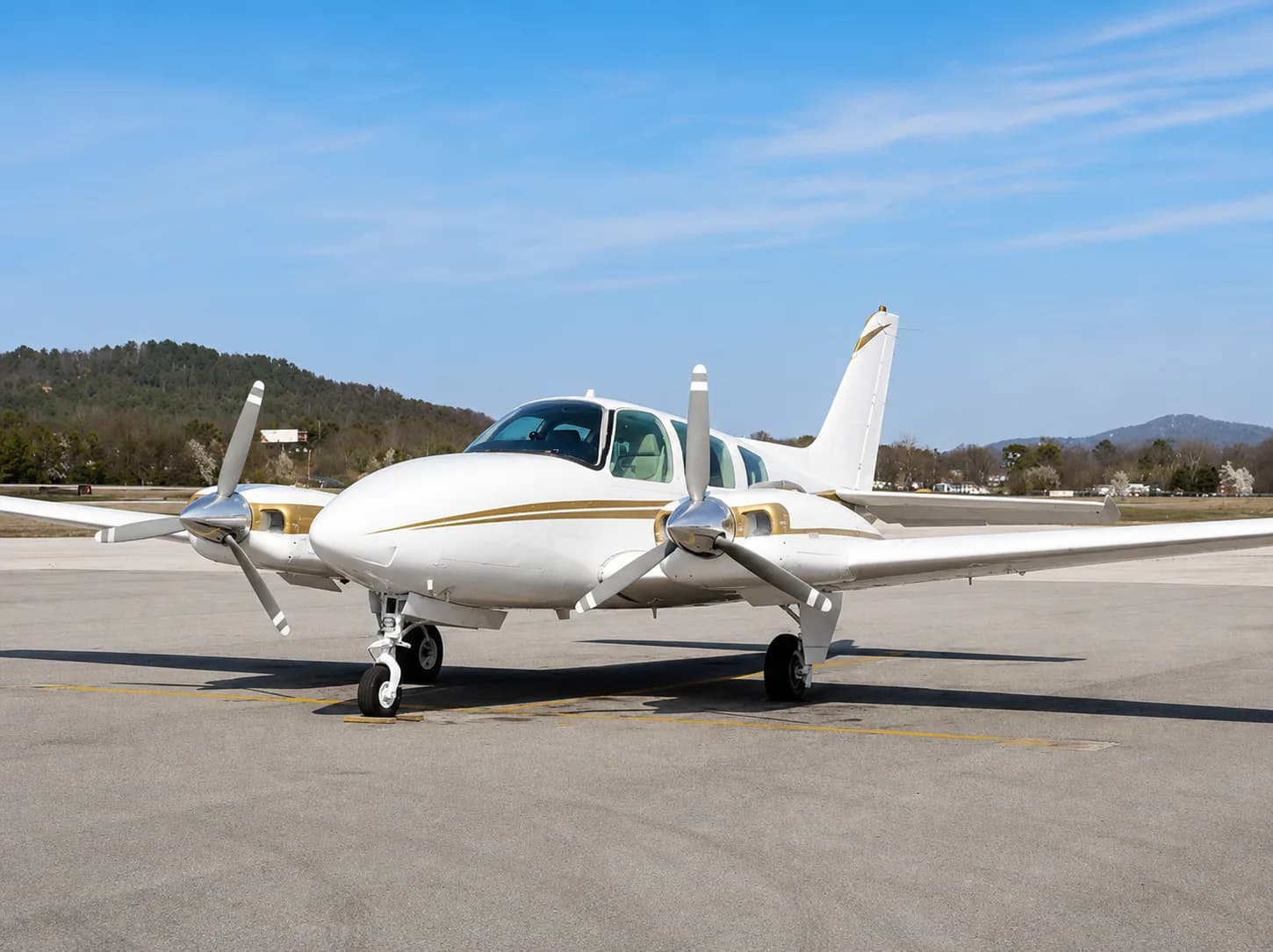 A white twin-engine propeller aircraft with gold stripes is parked on an asphalt runway under a clear blue sky.