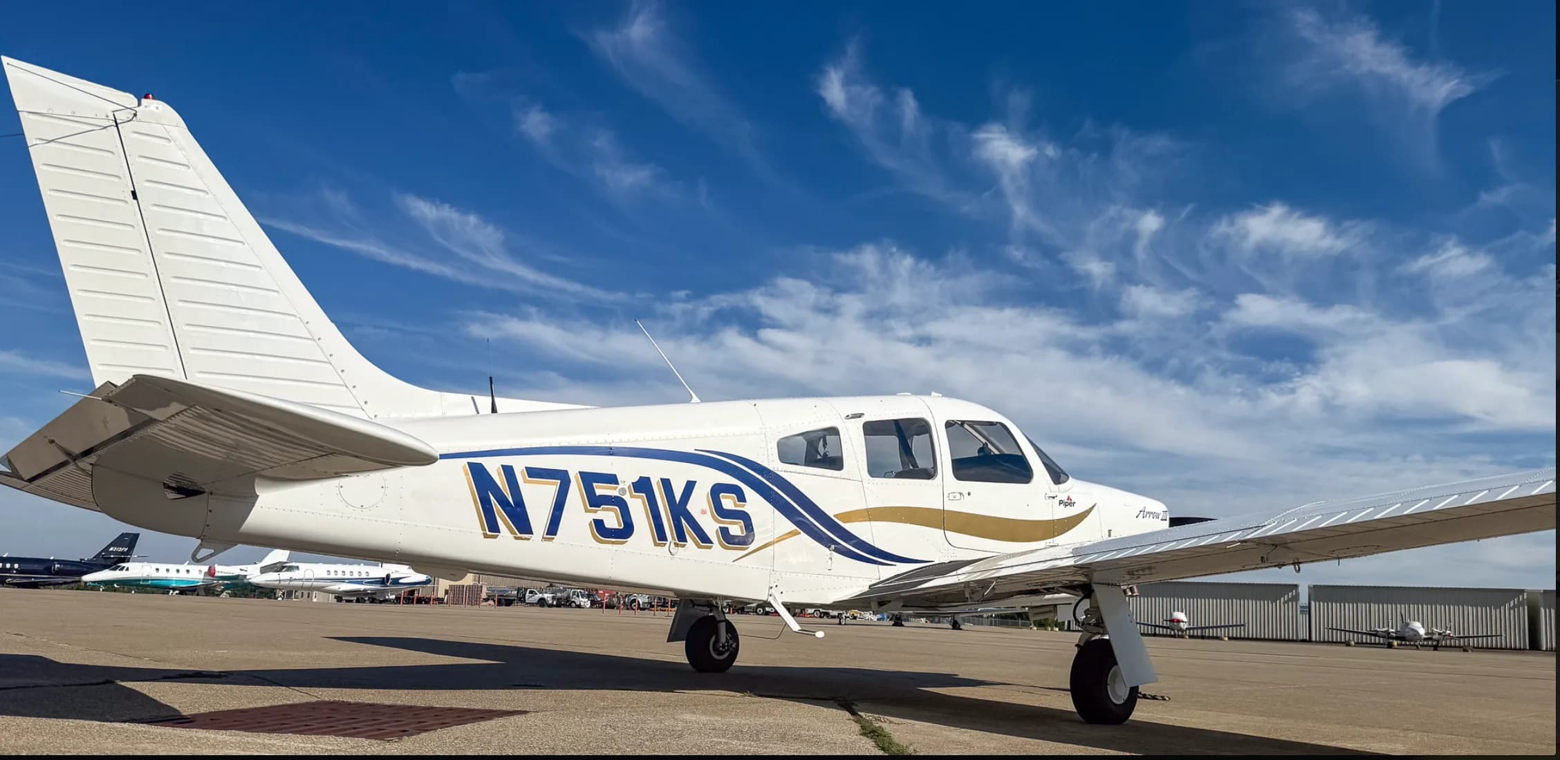 A white Piper Arrow III aircraft with blue and gold stripes, tail number N751KS, parked on an airport tarmac under a blue sky with wispy clouds.
