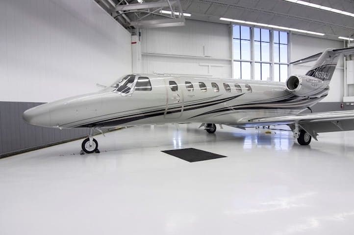 A silver Cessna Citation CJ3+ aircraft with black and grey stripes on the fuselage and tail, parked inside a brightly lit hangar.