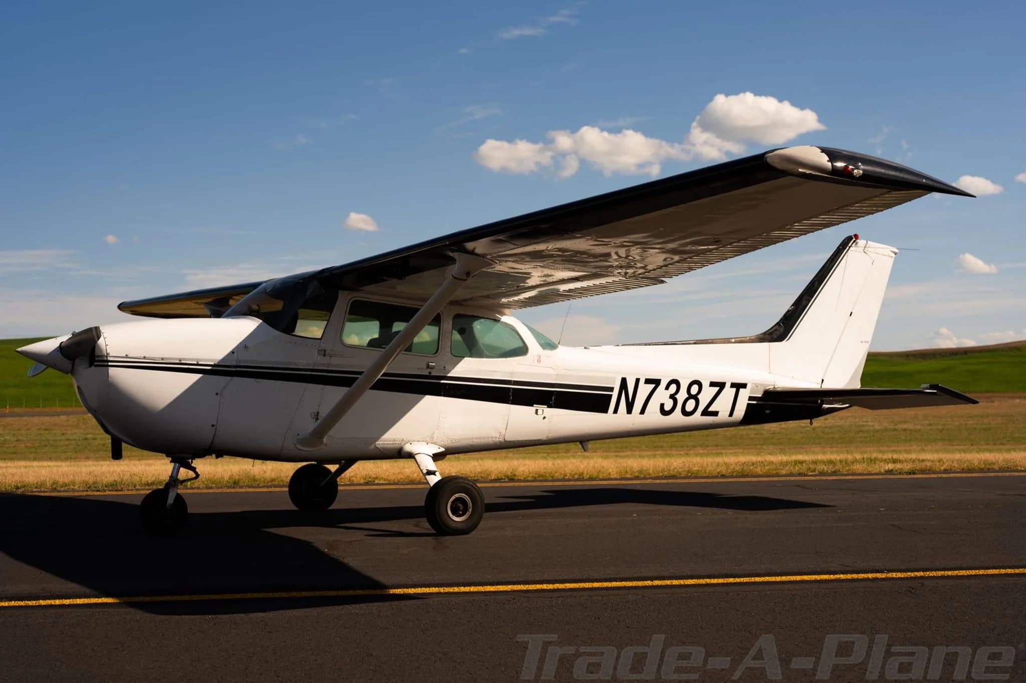 A white Cessna 172 with black stripes, tail number N738ZT, parked on an airfield under a partly cloudy sky.