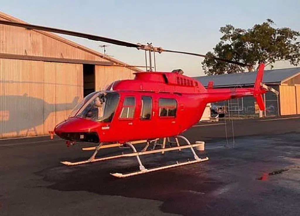 A red helicopter with white skids is parked on an asphalt apron in front of hangars at sunset.