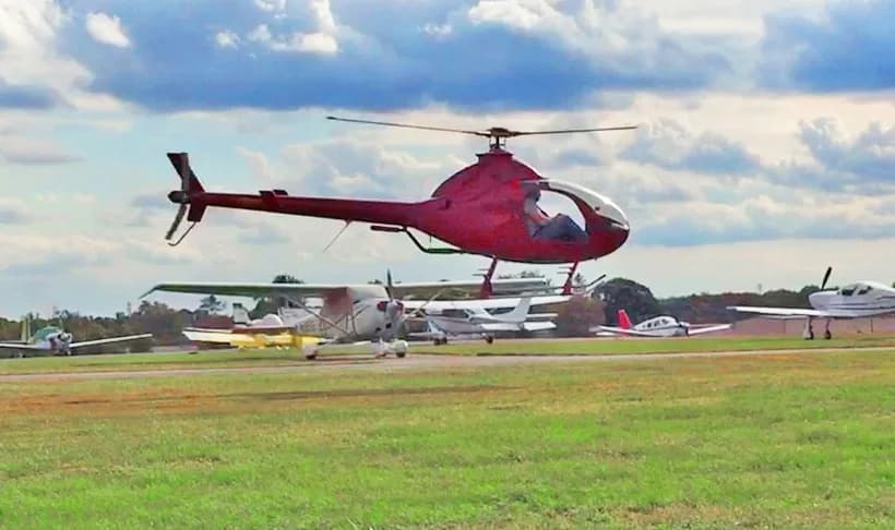A red RotorWay Exec helicopter hovers above an airfield with several small planes parked in the background.