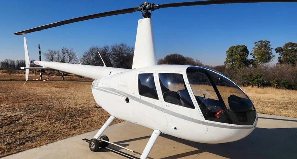 A white Robinson R44 helicopter with gray stripes is seen from the front-right, parked on a concrete pad.
