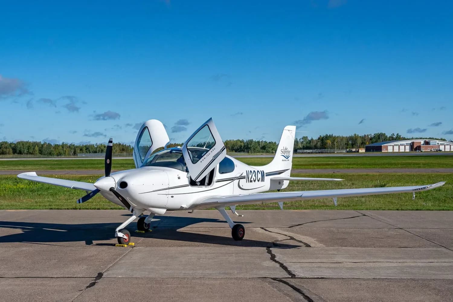 A white Cirrus SR20 aircraft with registration N23CM and "Lake Superior College" logo on the tail, parked on an airfield with doors open.