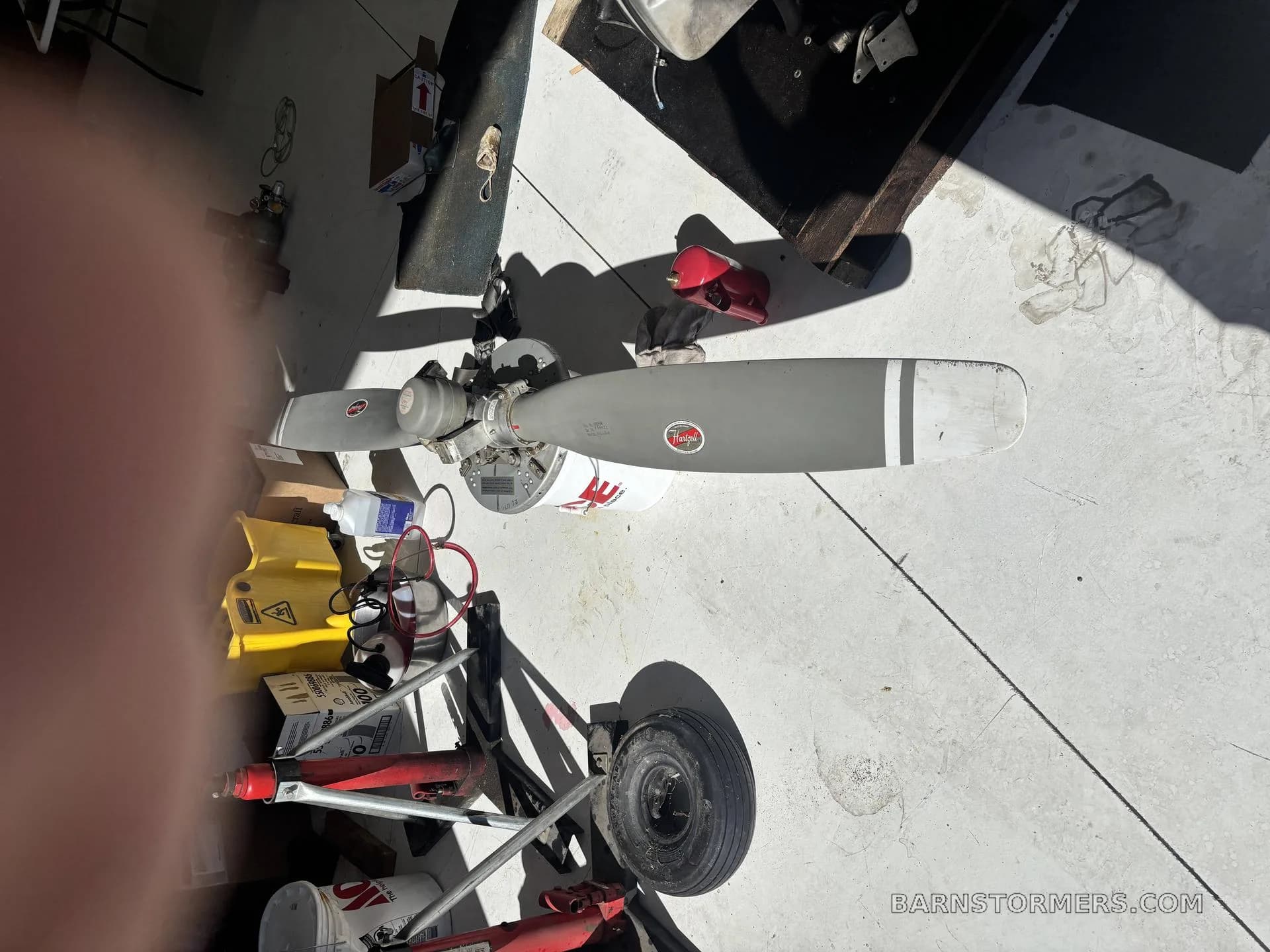 A two-bladed aircraft propeller with a red Hartzell logo, lying on a concrete floor surrounded by tools and equipment.