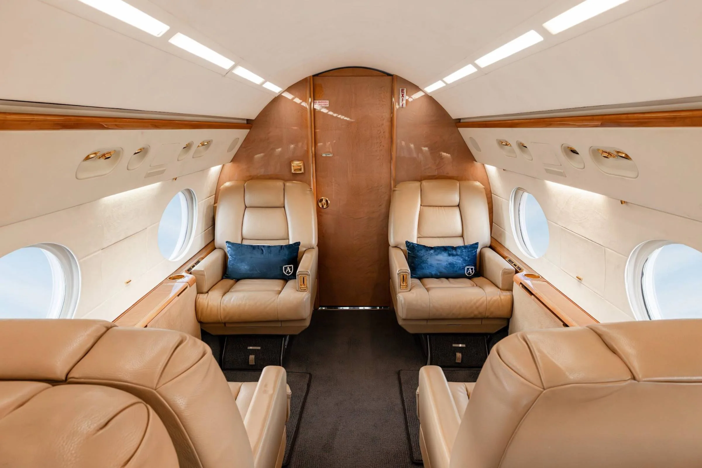 The forward section of a private jet cabin with tan leather seats and blue pillows, facing a wooden bulkhead.