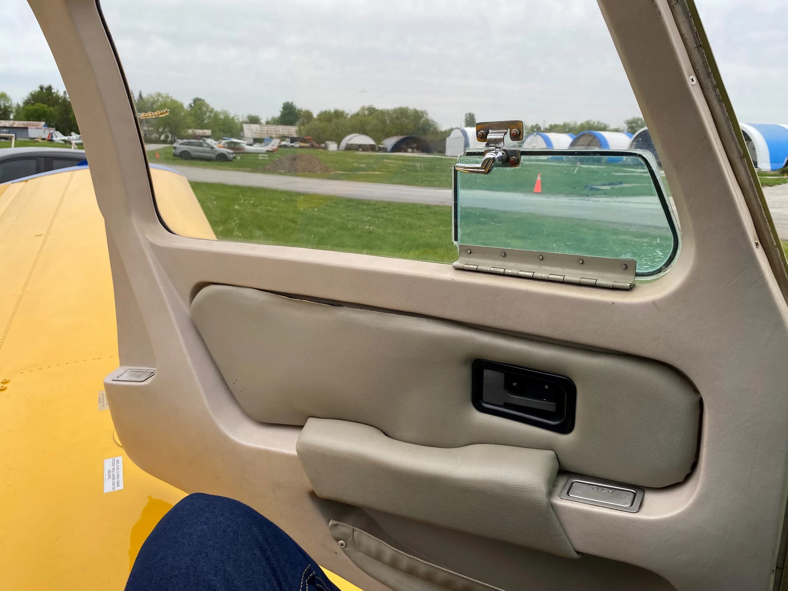 View from inside the aircraft cabin looking out the window at an airfield with hangars and other aircraft.