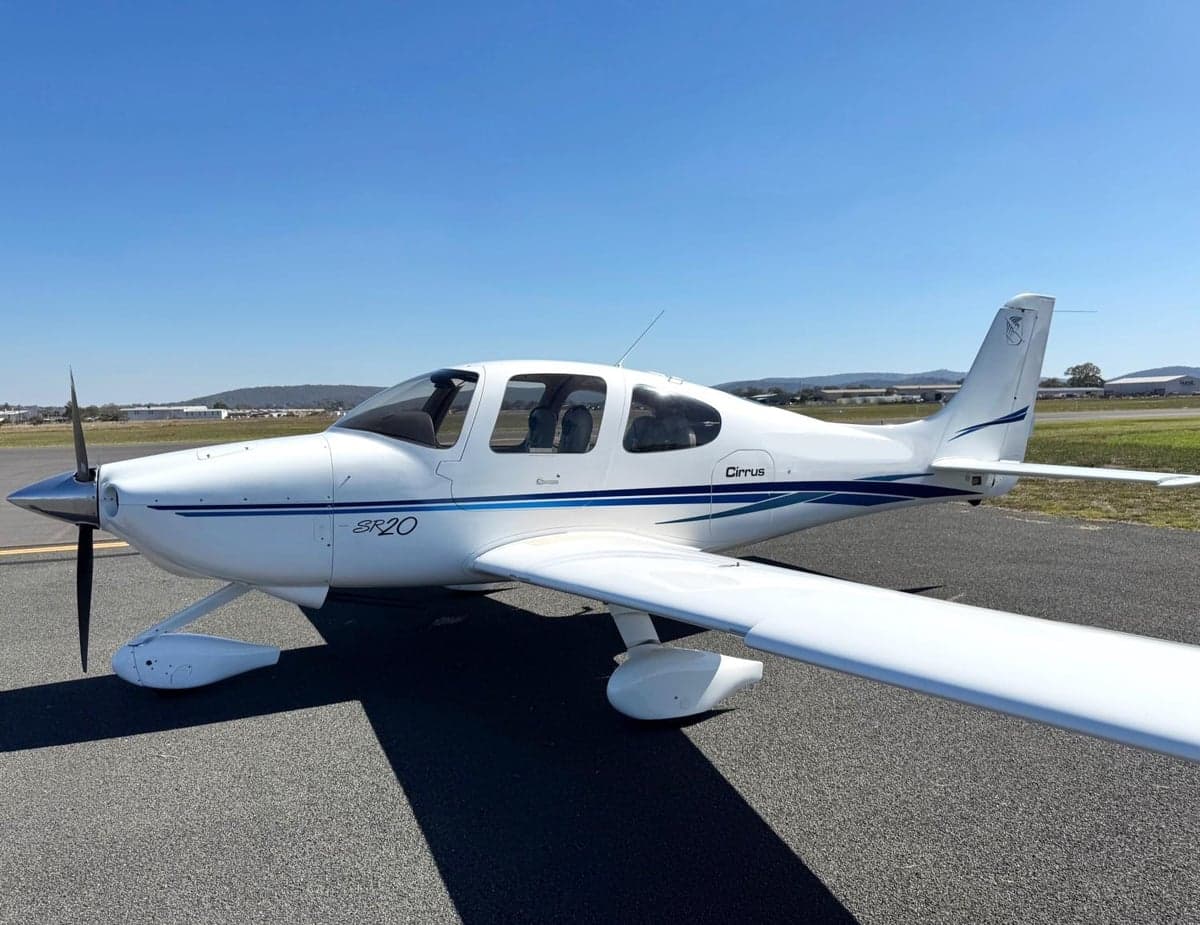 A white Cirrus SR20 aircraft with blue stripes, parked on an asphalt runway under a clear blue sky.