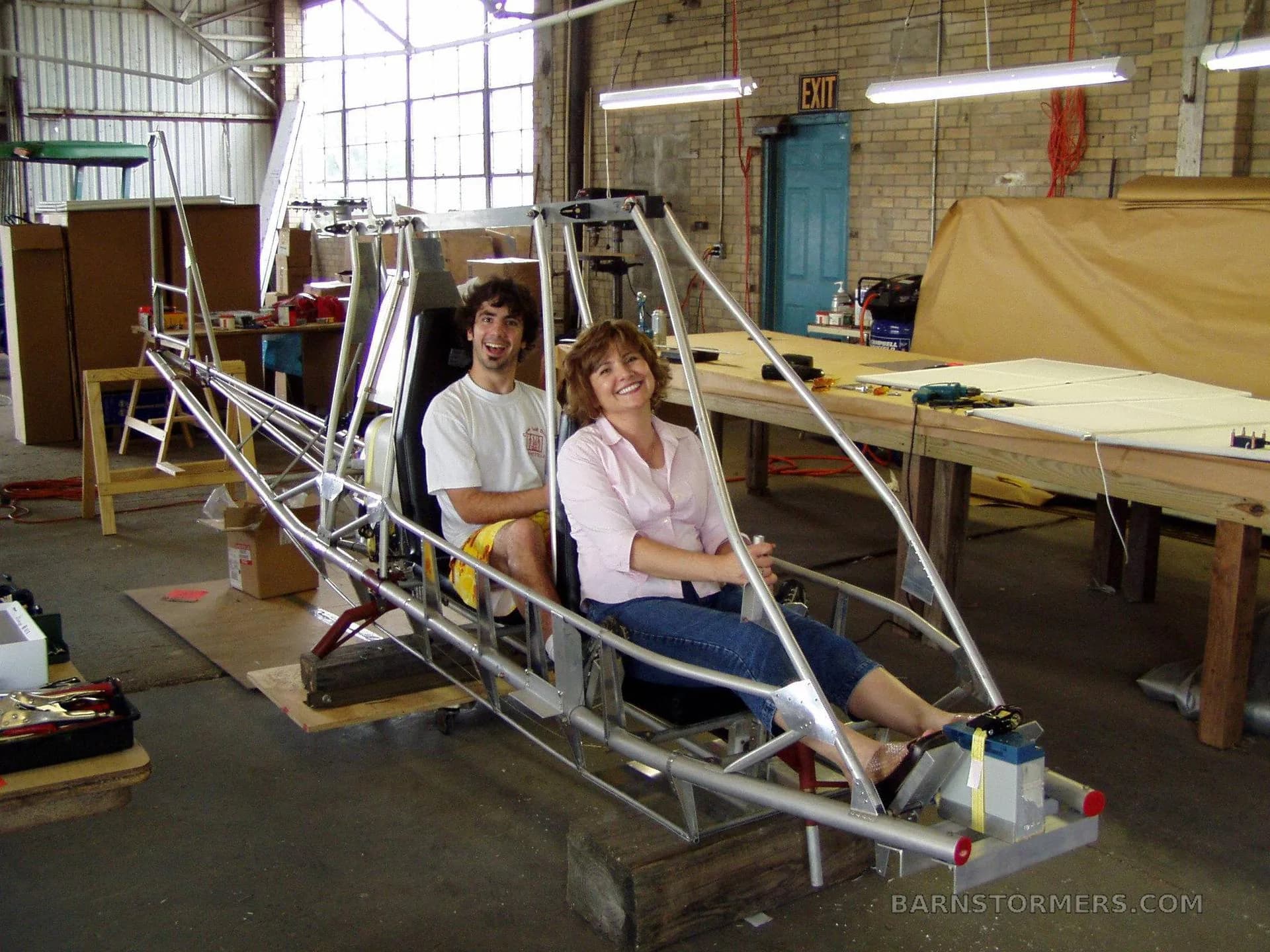 A man and a woman sitting in the metal frame of an experimental aircraft under construction in a workshop.