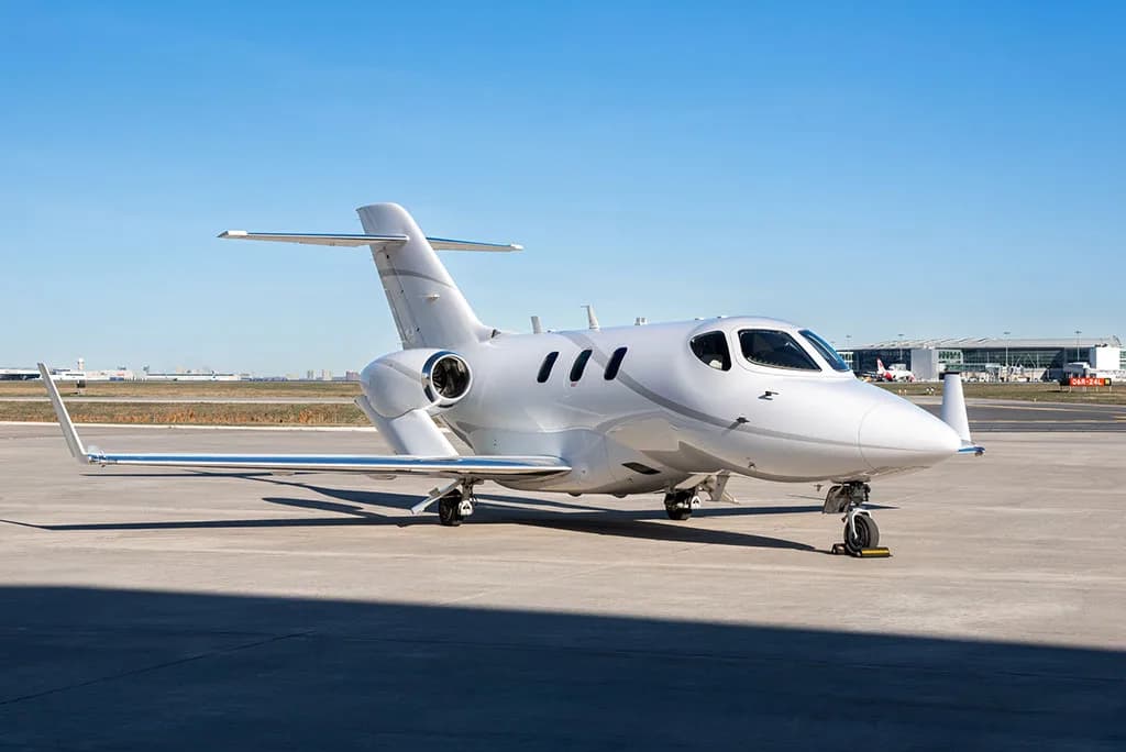 A white HondaJet HA-420 business jet parked on an airport tarmac with other aircraft and buildings in the distance.