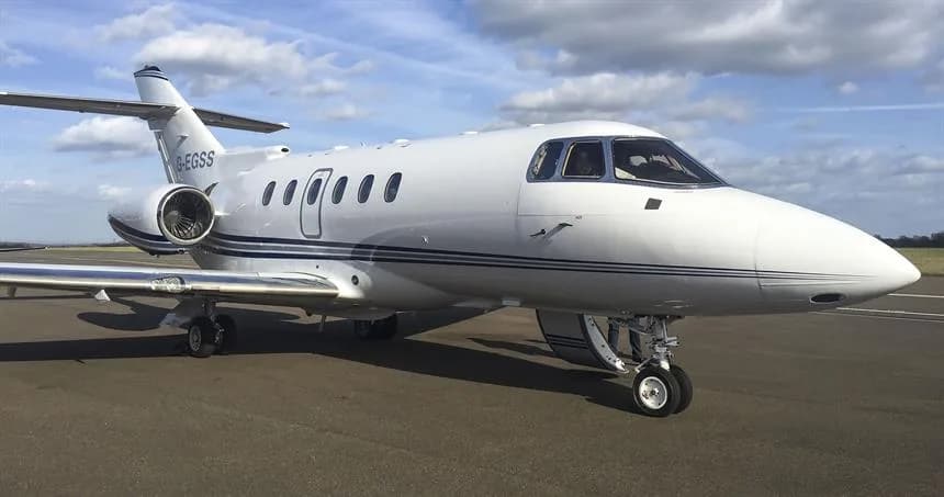 A white business jet with blue stripes, registration G-EGSS, parked on an airfield under a partly cloudy sky.