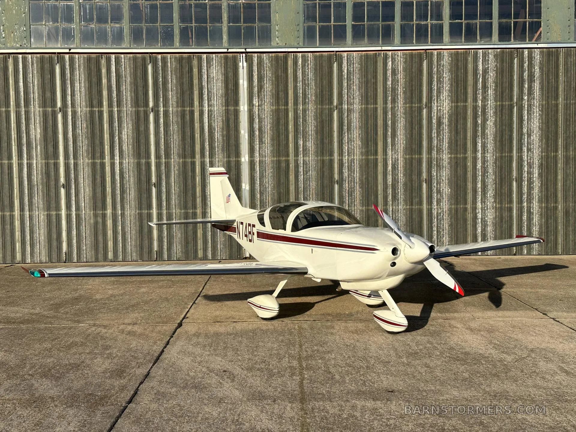 A white single-engine aircraft with a maroon stripe and registration N74BF is parked on concrete, viewed from the front-right, in front of a hangar.