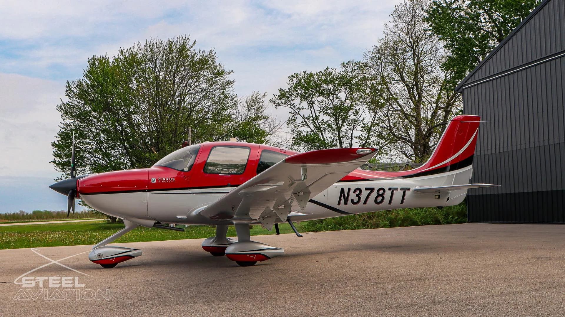A red and white Cirrus SR22 aircraft with tail number N378TT is parked on an apron, viewed from the right side with trees in the background.