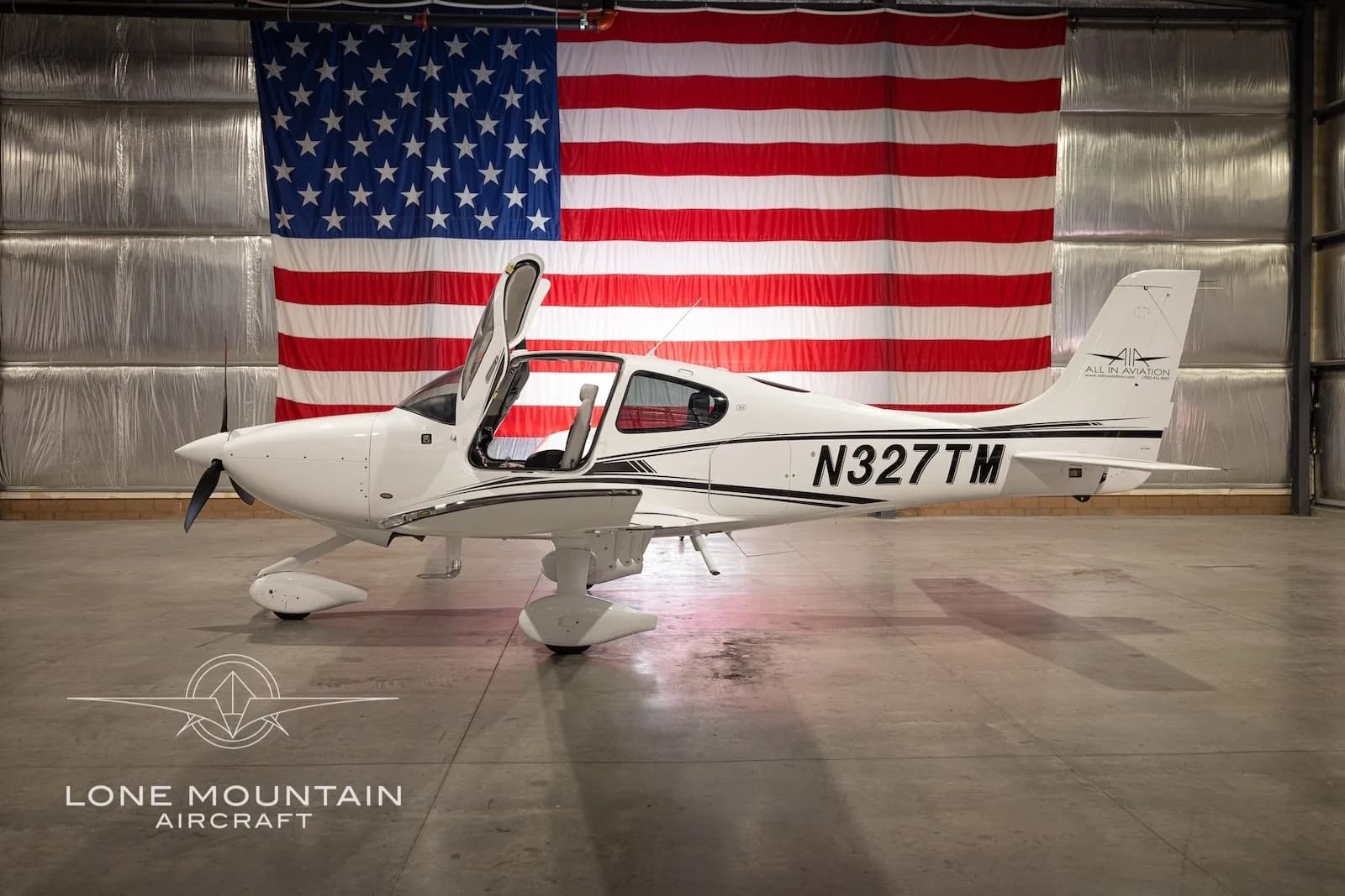 A white Cirrus SR22 aircraft with black stripes and registration N327TM, parked in a hangar with an American flag backdrop, with its gull-wing doors open.