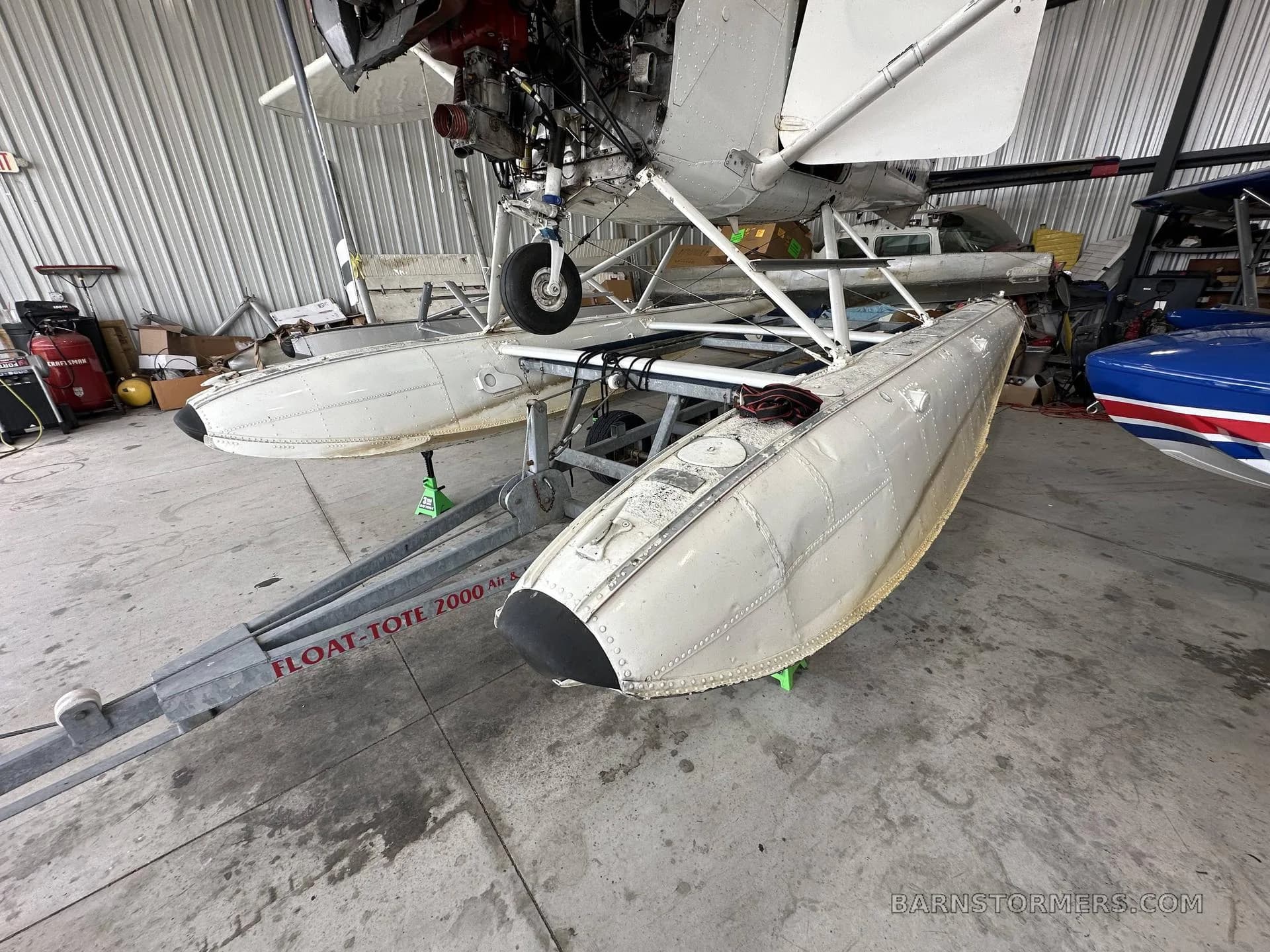 A white floatplane is shown from a slightly different angle in a hangar, highlighting its floats and exposed engine.