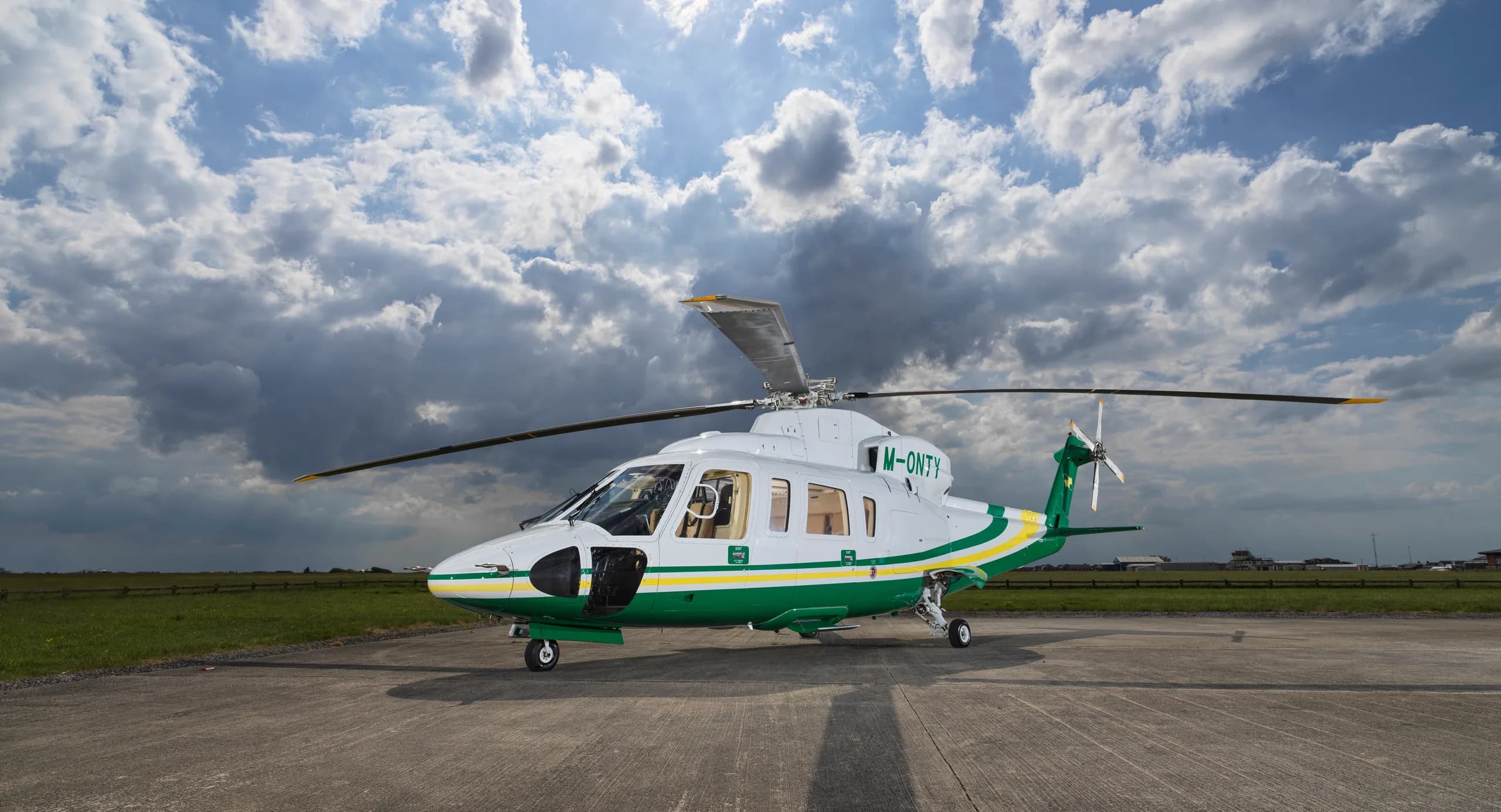 A white and green helicopter with registration M-ONTY sits on a tarmac under a dramatic cloudy sky.
