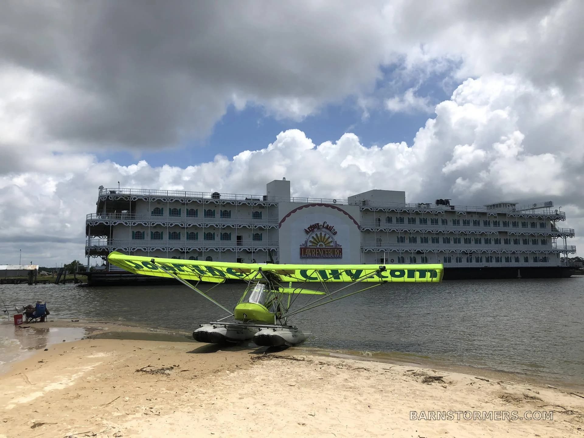 A bright green ultralight seaplane with "Soar The City.com" on its wings rests on a sandy shore next to a large white riverboat under a cloudy sky.