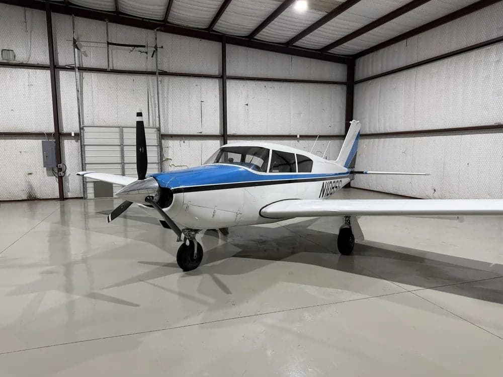 A white and blue Piper PA-28-140 Cherokee is parked in a hangar.