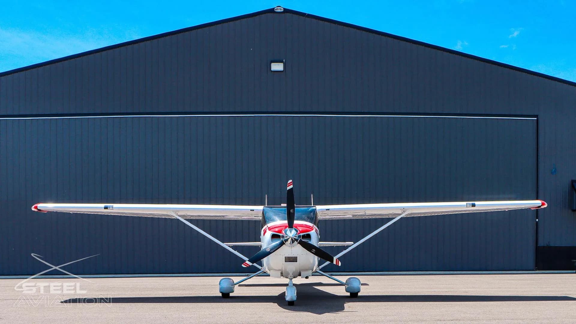 A white Cessna T182T Skylane TC with red and grey stripes is parked head-on in front of a large dark hangar under a clear blue sky.