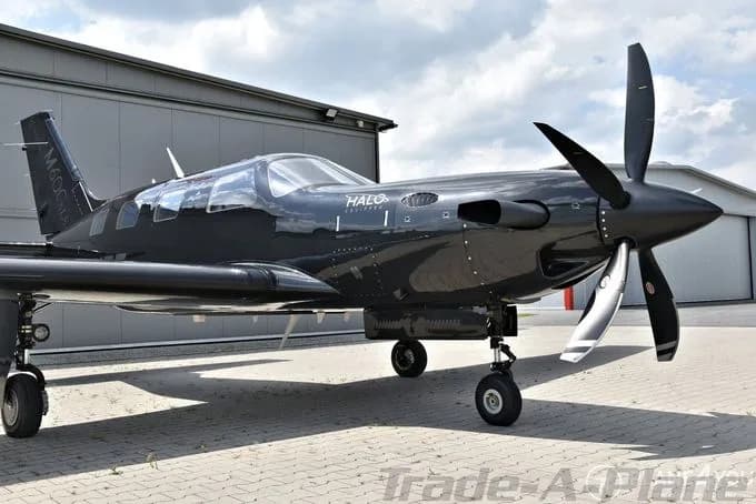 A black Piper M600/SLS 'HALO' edition turboprop aircraft with a three-blade propeller, parked on an apron next to a hangar.