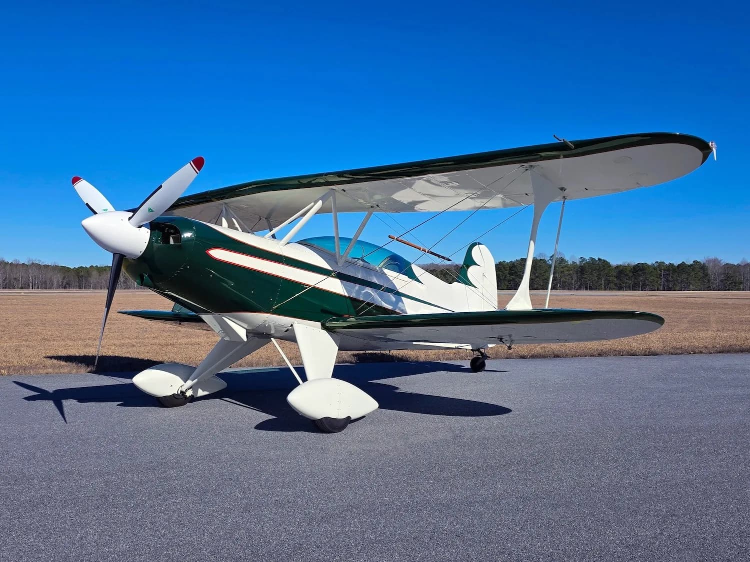 A green and white biplane with a red stripe sits on an asphalt runway under a clear blue sky.