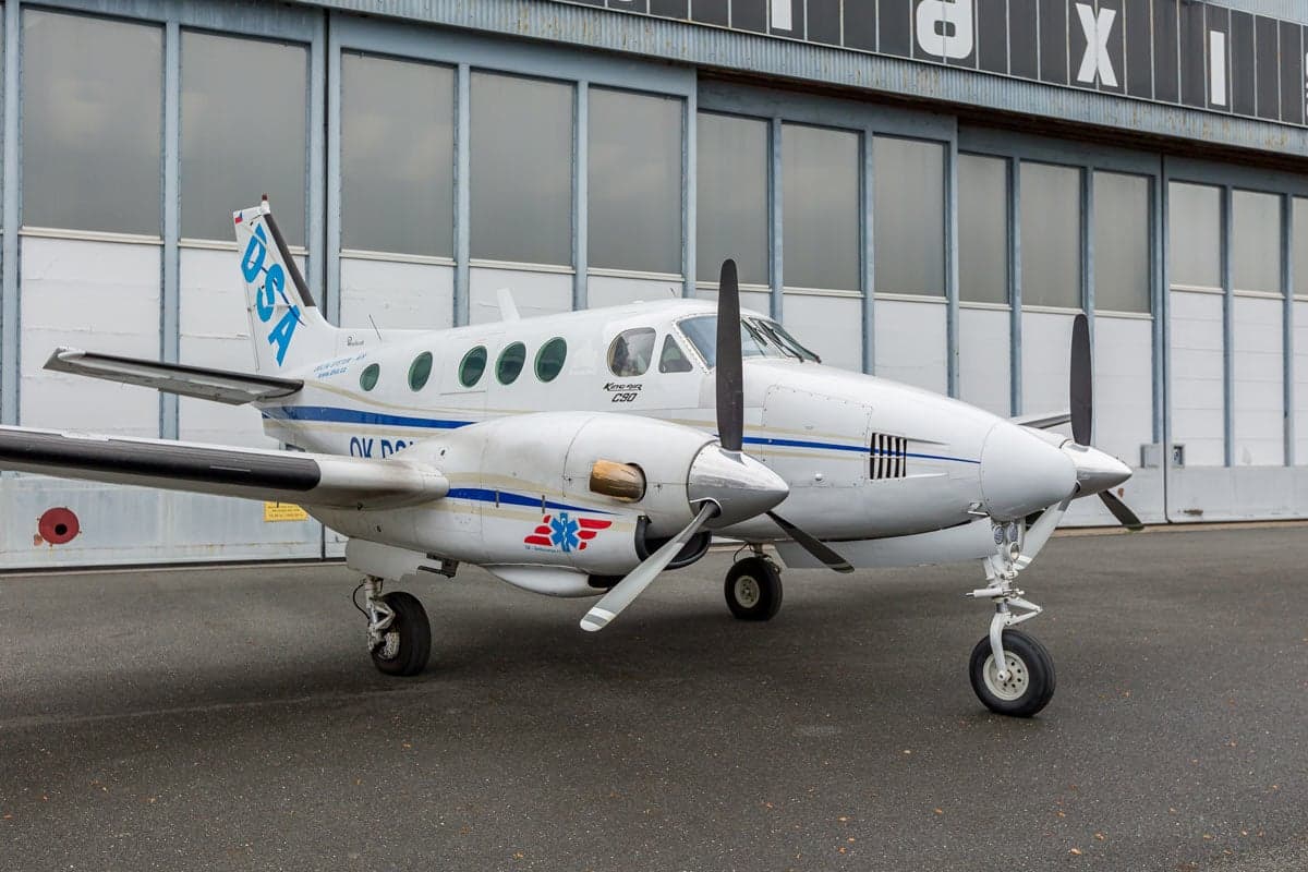 A Beechcraft King Air C90 aircraft with a white and blue paint scheme, parked in front of a hangar.