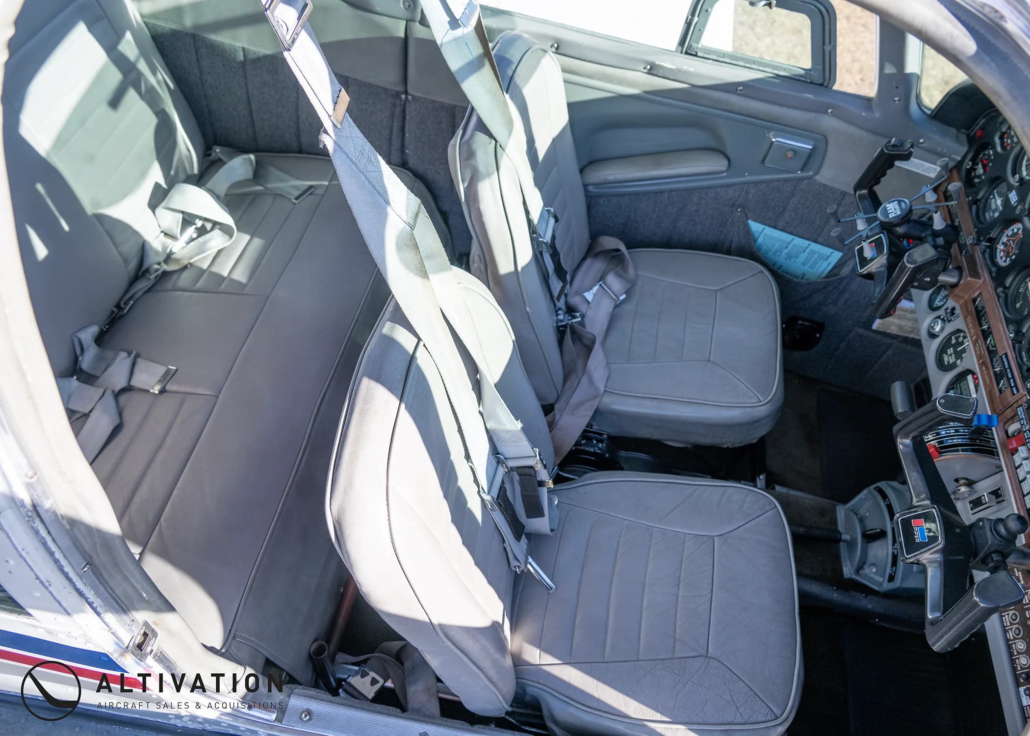 The gray interior cabin of a small aircraft, showing the front and rear seats with seatbelts.