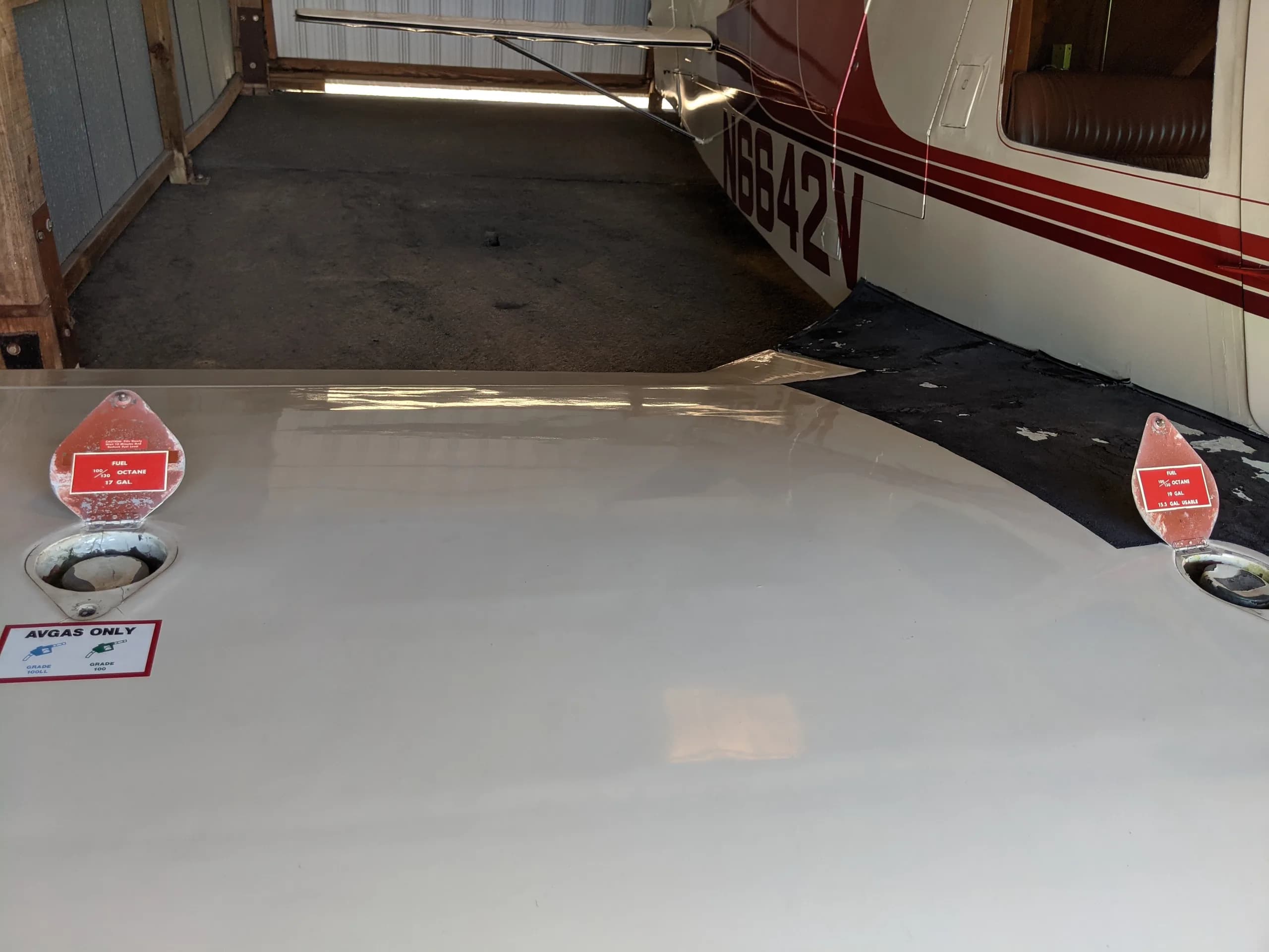 A close-up view of the white wing surface with two red fuel caps and "AVGAS ONLY" label, next to the aircraft fuselage.