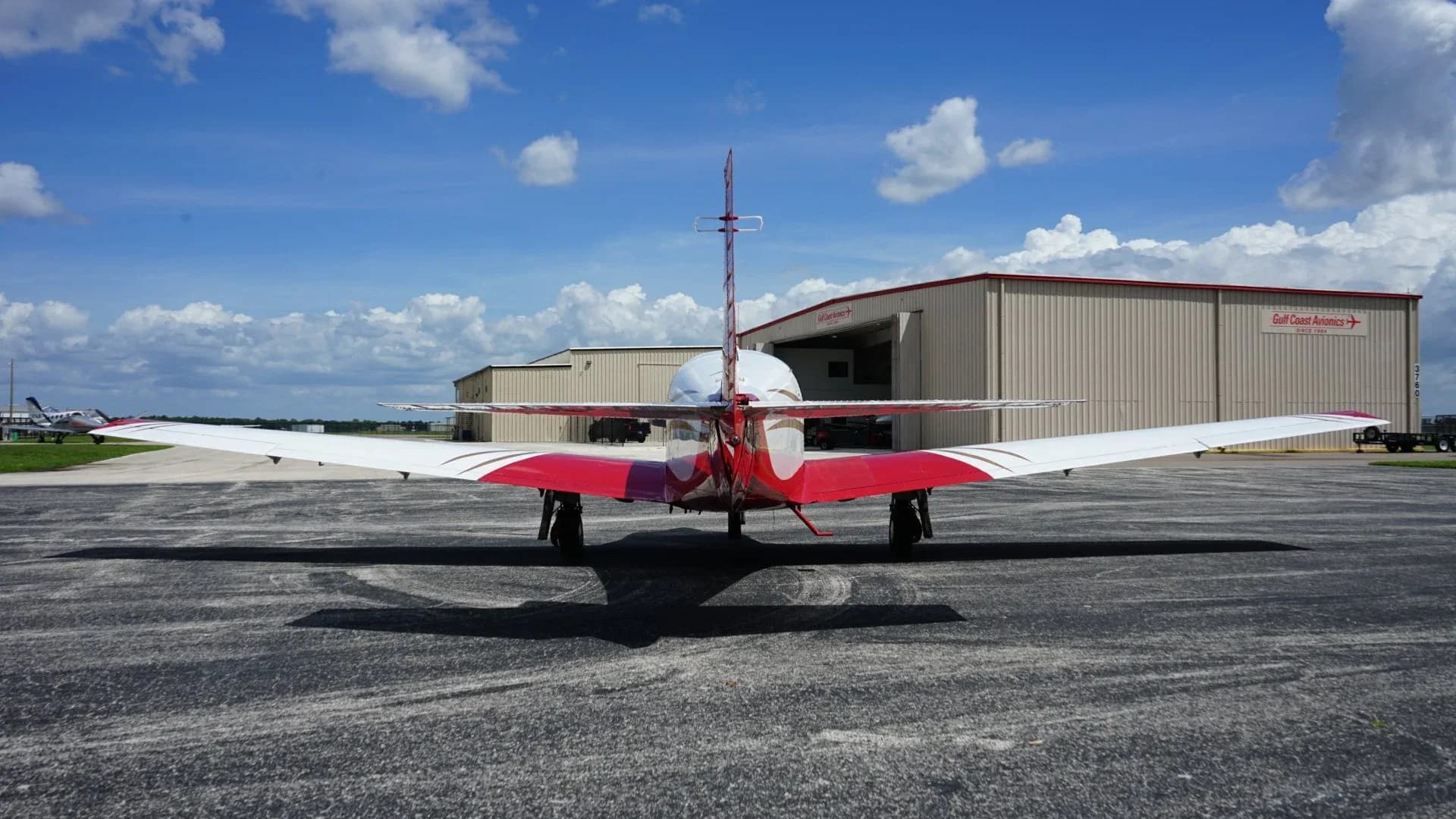 A rear view of a red and white single-engine aircraft with gold stripes parked on an asphalt apron under a blue sky.