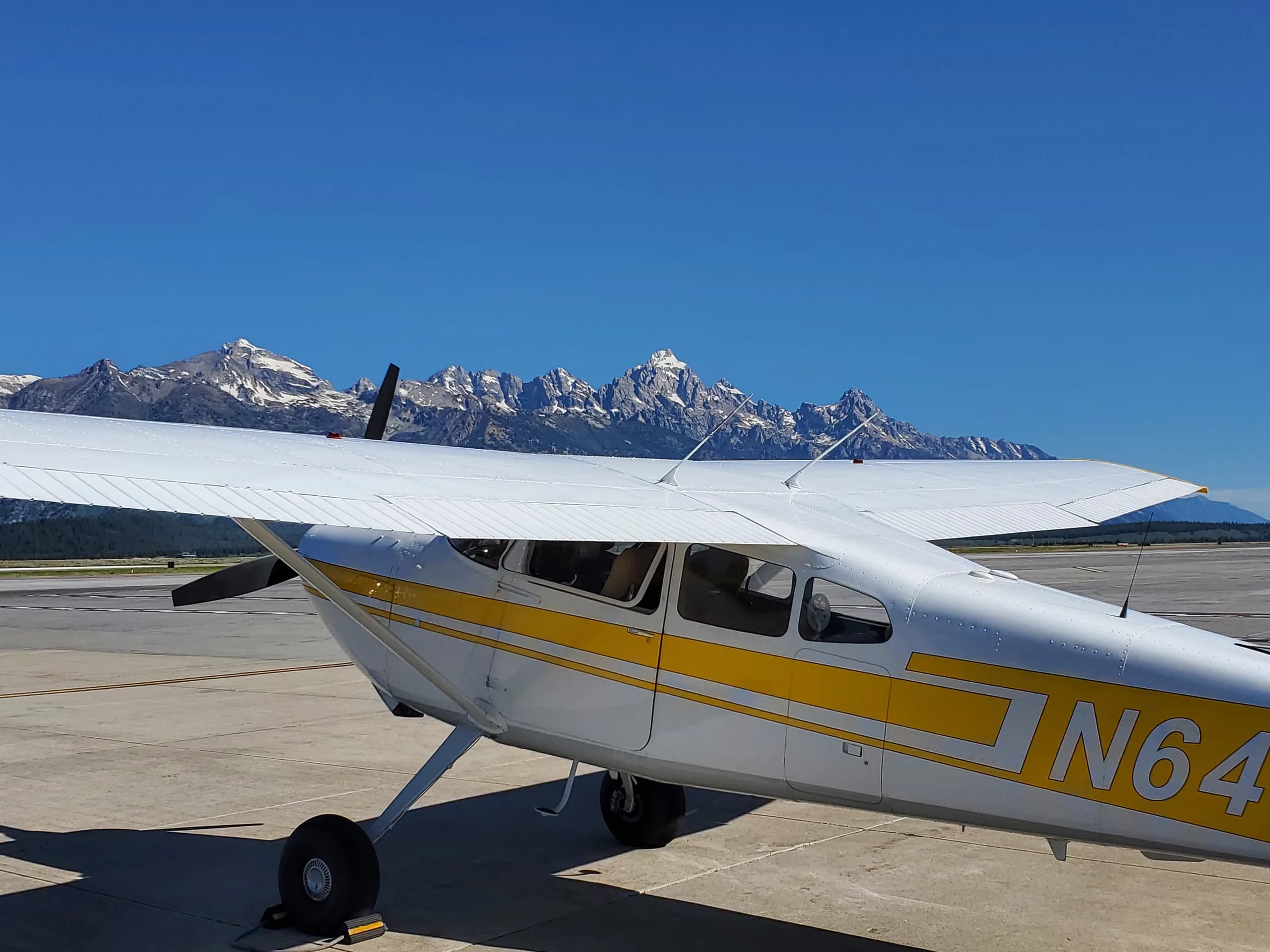 A white and yellow Cessna 172 aircraft with registration N644FM parked on an airfield with snow-capped mountains in the background.