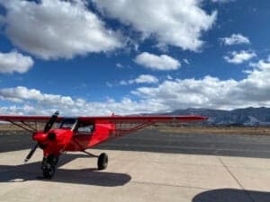 A red Cubcrafters aircraft is parked on a runway with mountains in the background.