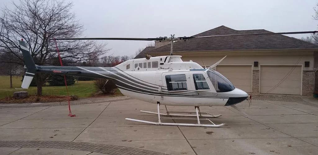 Side view of a white helicopter with dark grey stripes parked on a concrete driveway next to a house.