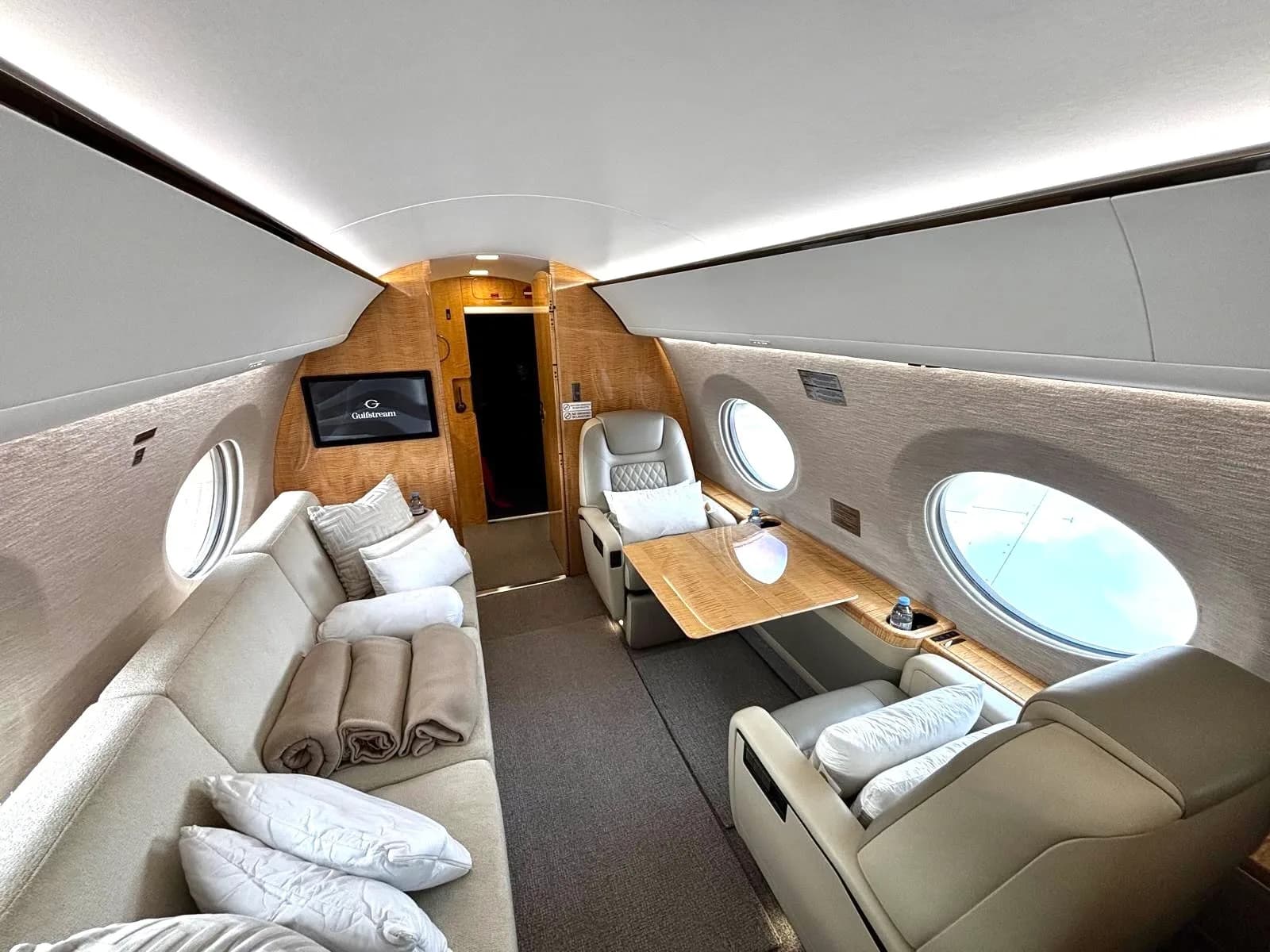 A luxurious private jet cabin featuring a cream-colored sofa with pillows and blankets, facing two individual seats with a table.