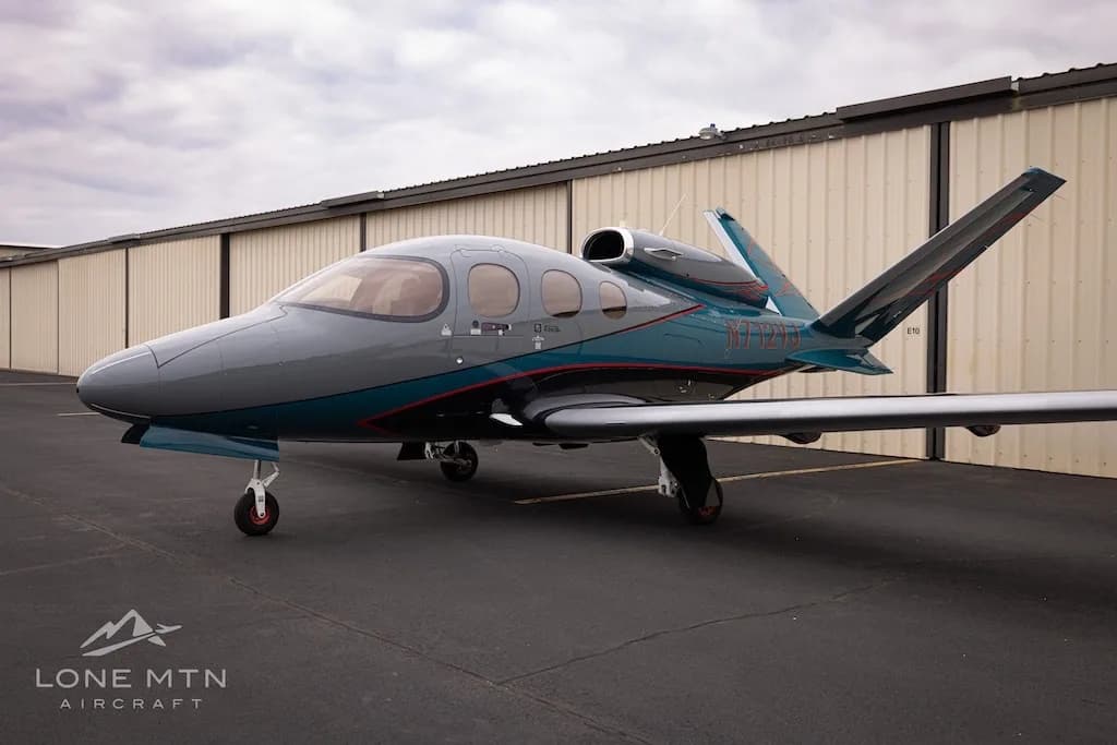 A Cirrus Vision Jet (N712VJ) with a grey and teal paint scheme, parked on an airfield next to hangars.