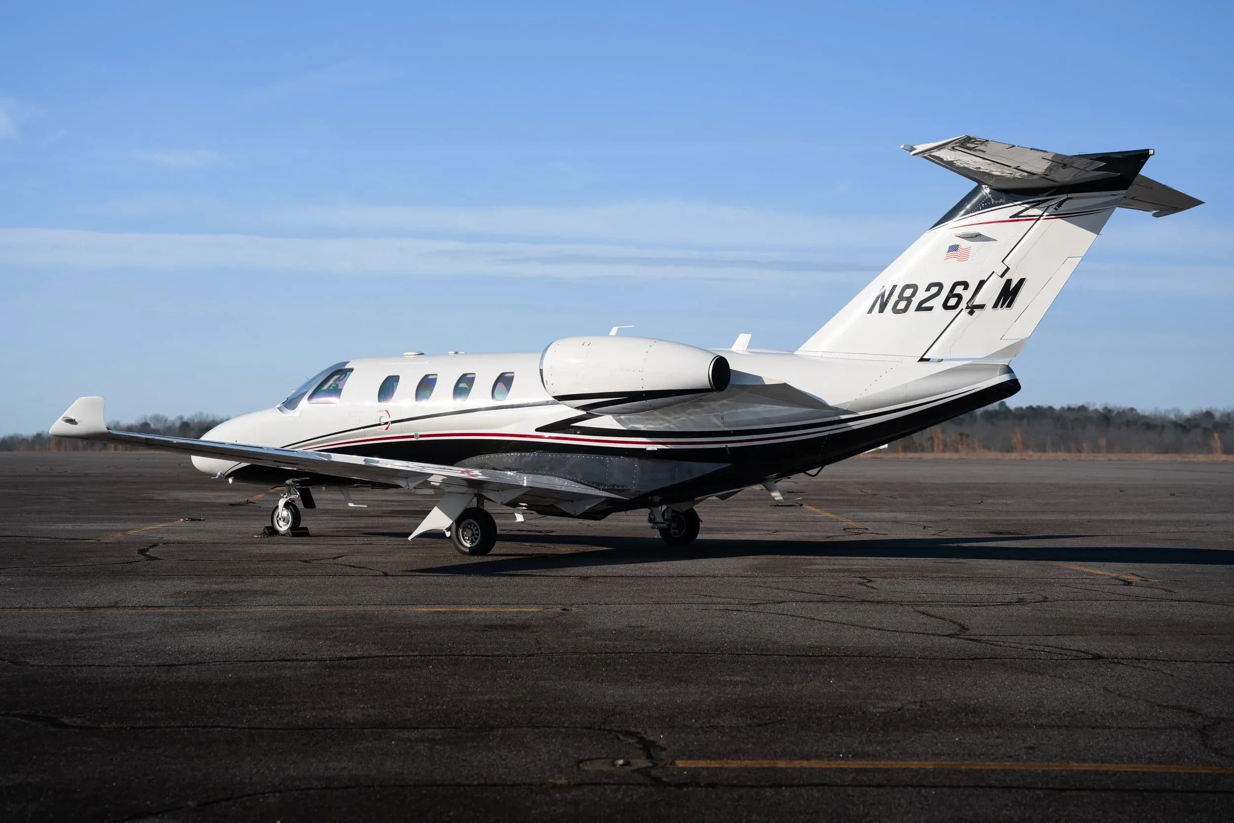 A white business jet with black and red stripes, registration N826LM, parked on an asphalt runway under a clear blue sky.