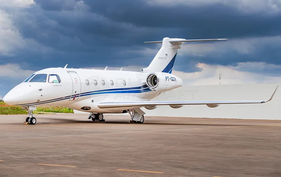A white Embraer Legacy 450 business jet with blue stripes is parked on an asphalt tarmac under a cloudy sky.