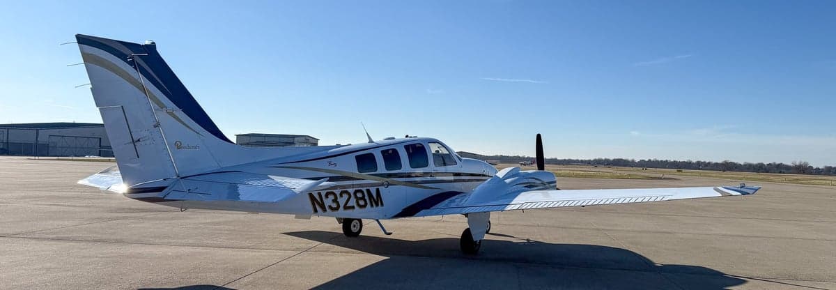 Rear-left view of a white twin-engine propeller aircraft N328M with black and gold stripes on an airfield.