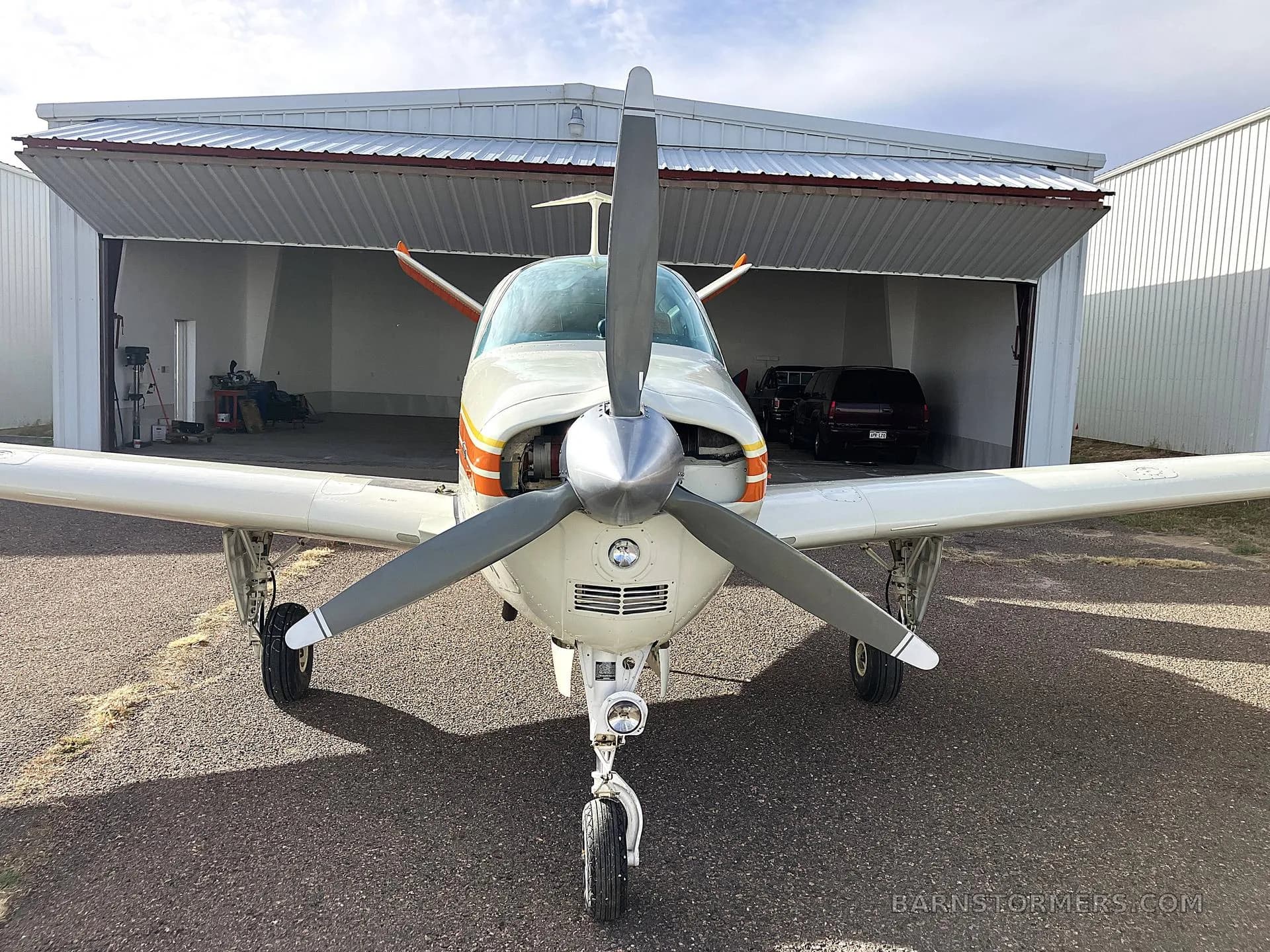 A head-on view of a white, orange, and yellow Beechcraft Bonanza N725AP with its propeller centered, parked in front of an open hangar.