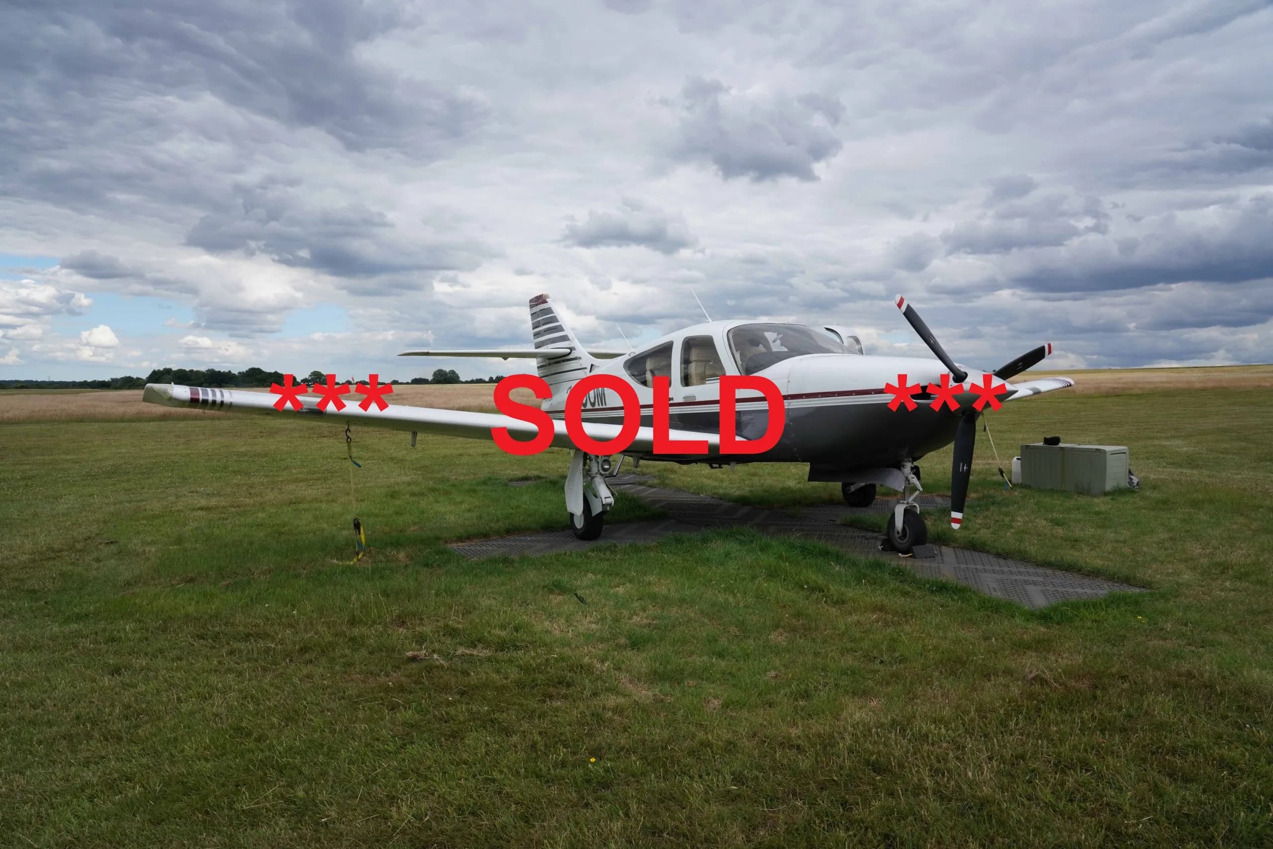 A white and grey single-engine propeller aircraft is parked on a grassy field under a cloudy sky.